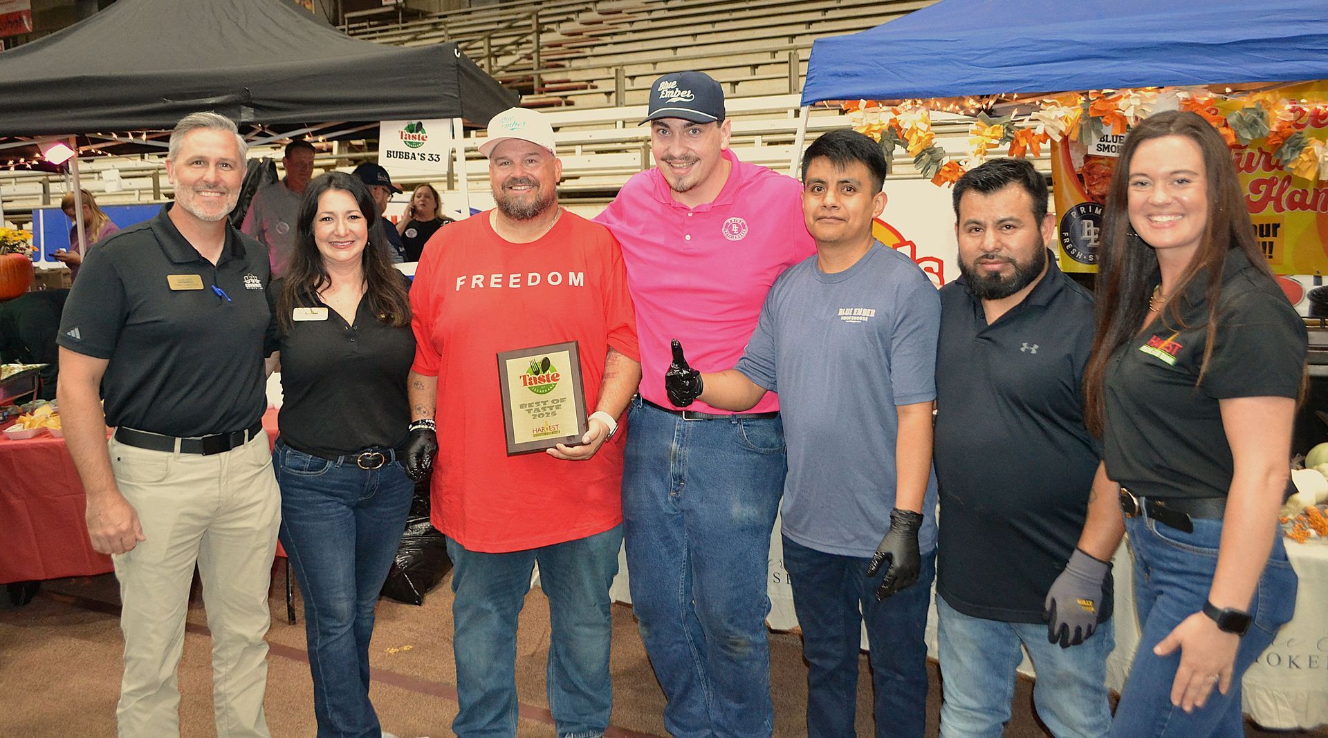 Seven people posing at an indoor event, one holding a small trophy plaque, with a pink banner and tents behind them