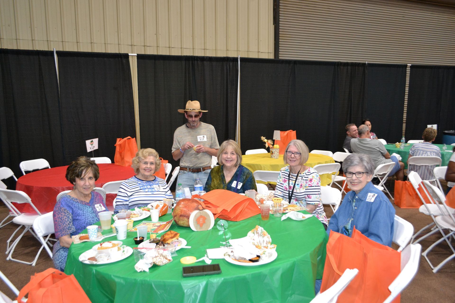 People seated around a green table at a banquet or community event, with orange bags and a black curtain backdrop