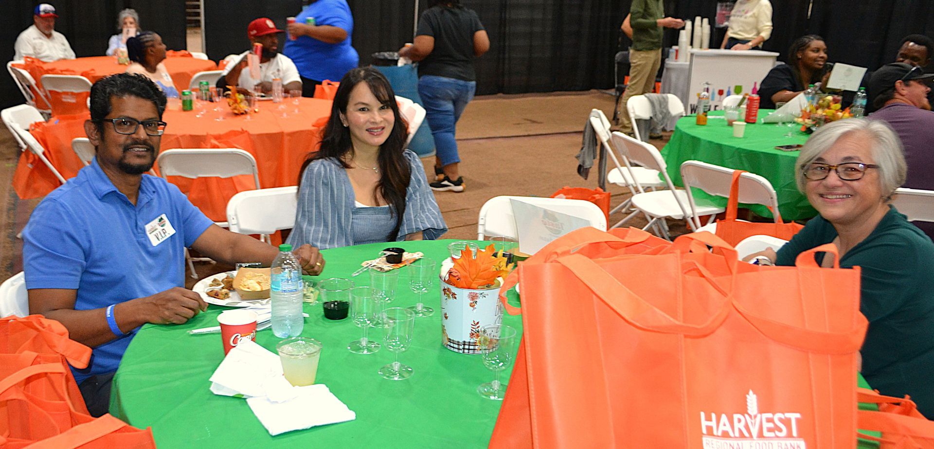 Three people seated at a green table at a community event with orange Harvest bags nearby