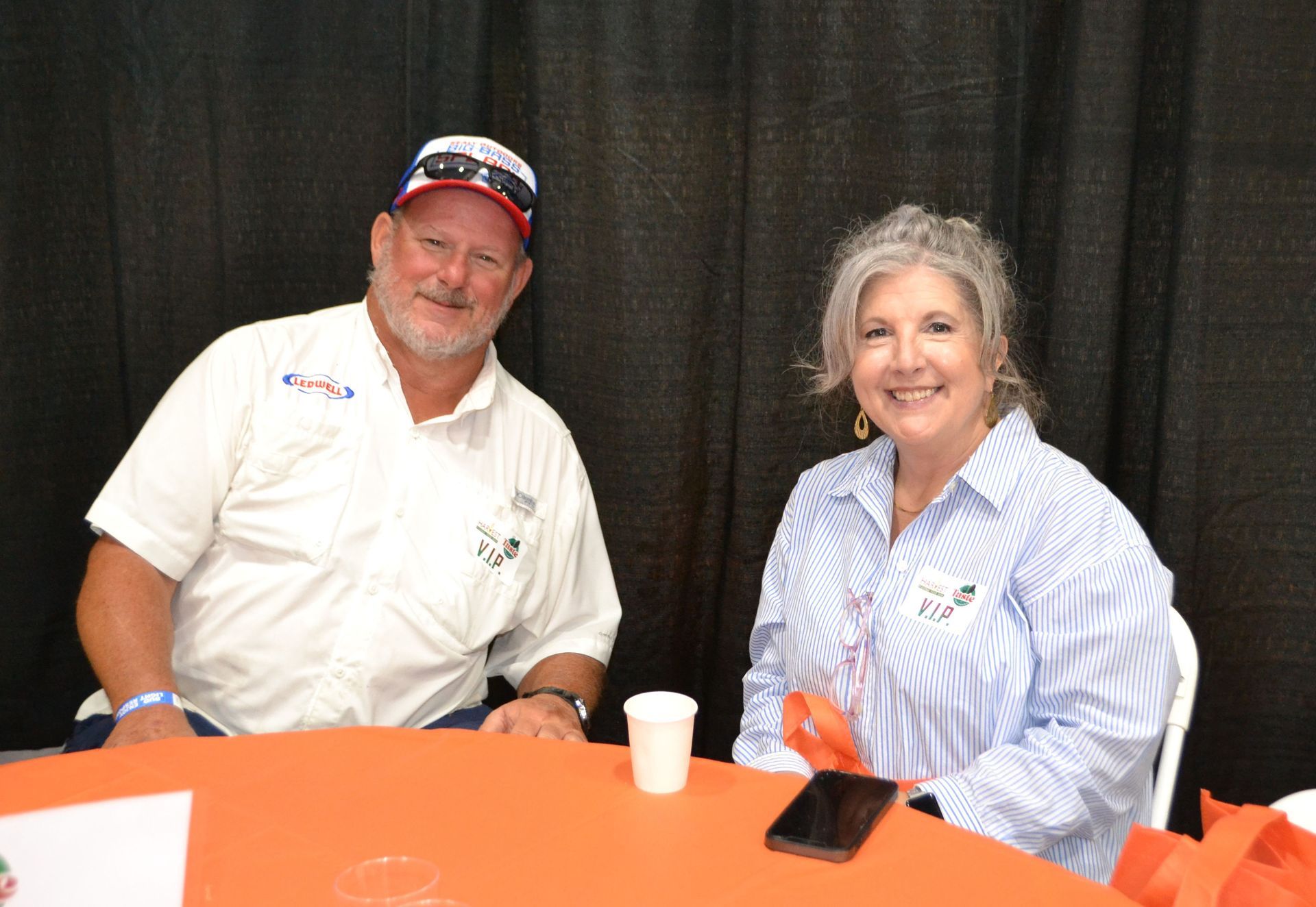 Two smiling people seated at an orange table with a white cup and phone in front of a black backdrop.