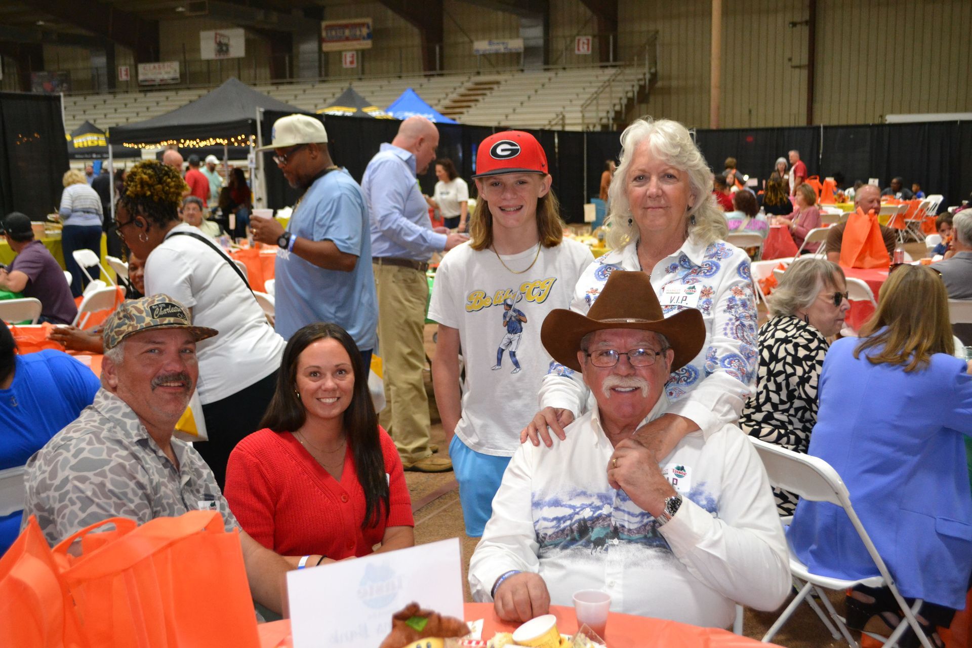 People seated at a crowded indoor event, posing at a table with bright tablecloths and orange decor.