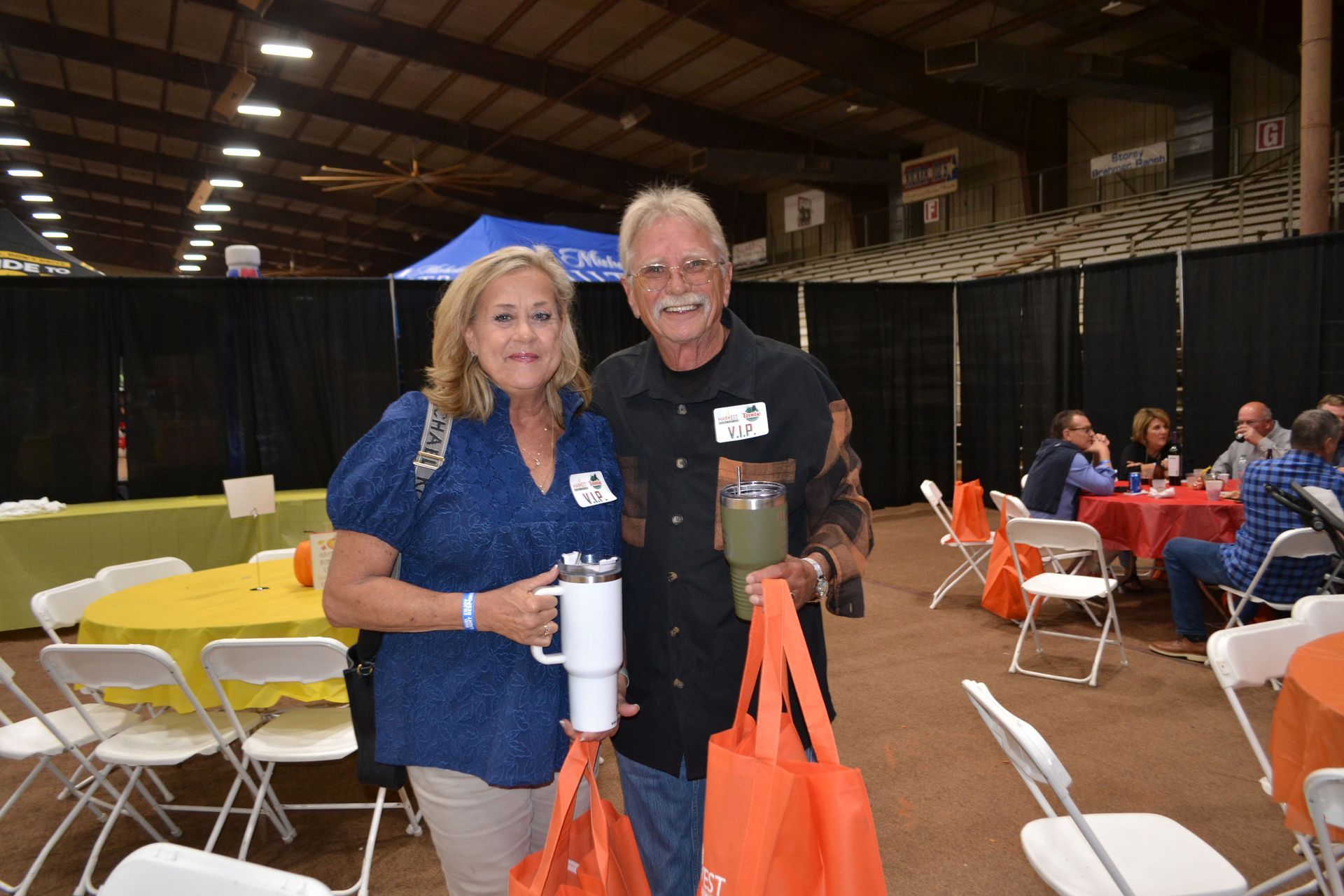 Two people smiling at an indoor event, holding drinks and orange bags near folding tables and chairs.