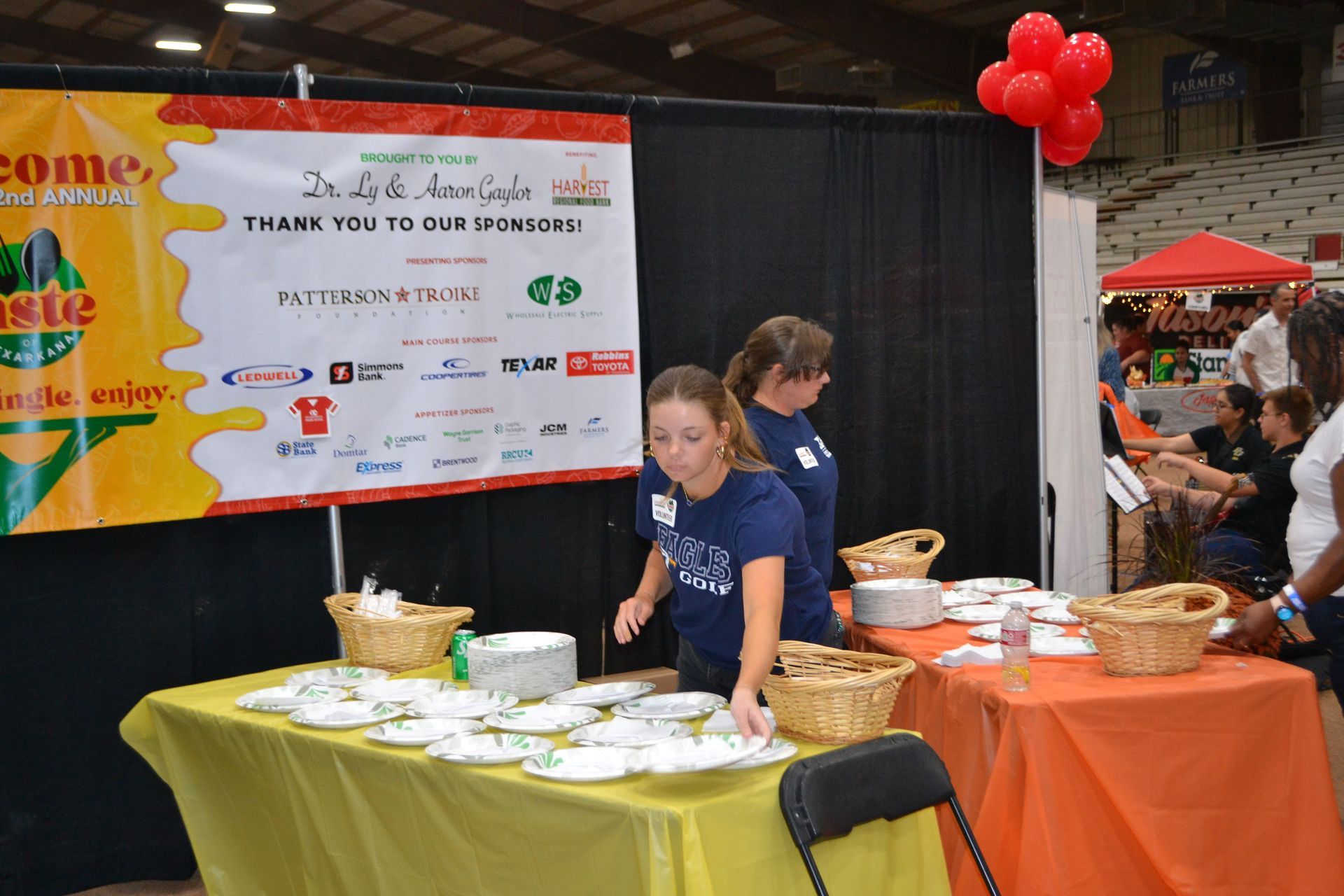 Two people serving food at a booth with yellow and orange tablecloths, signs, and red balloons.