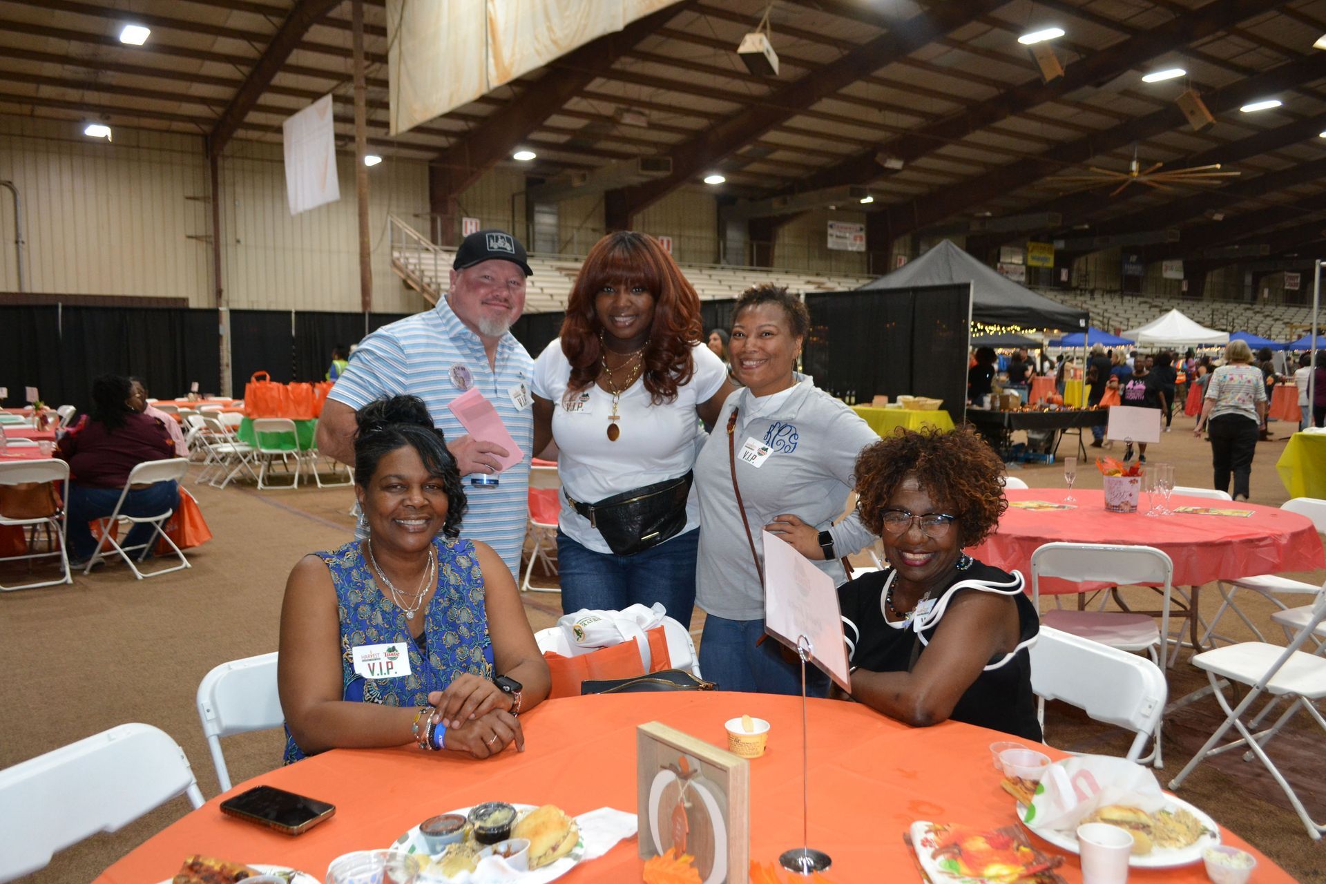 Five people posing at a table in a large indoor event hall with orange tablecloths.