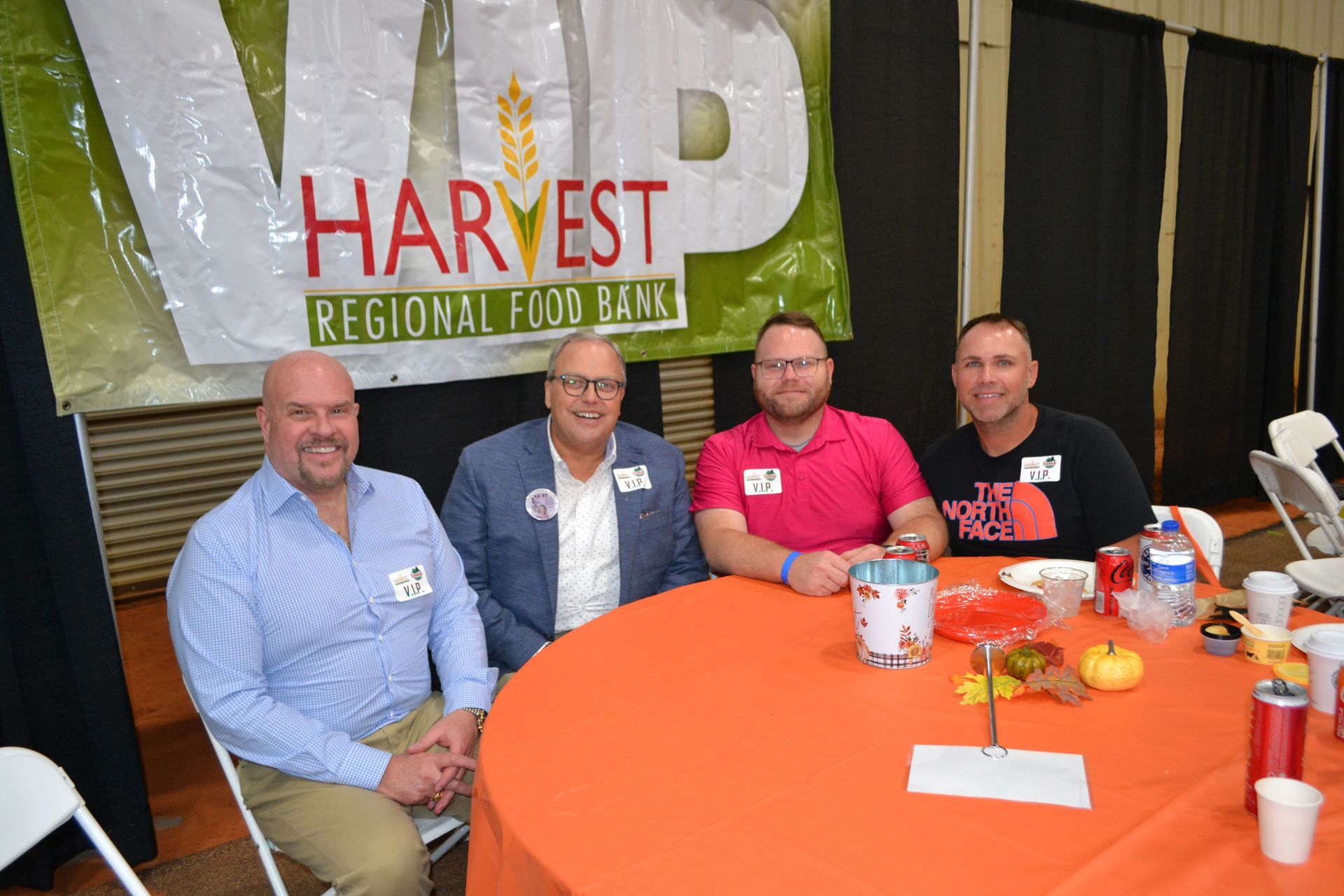 Four men seated at a round orange table at a Harvest Regional Food Bank booth, smiling indoors