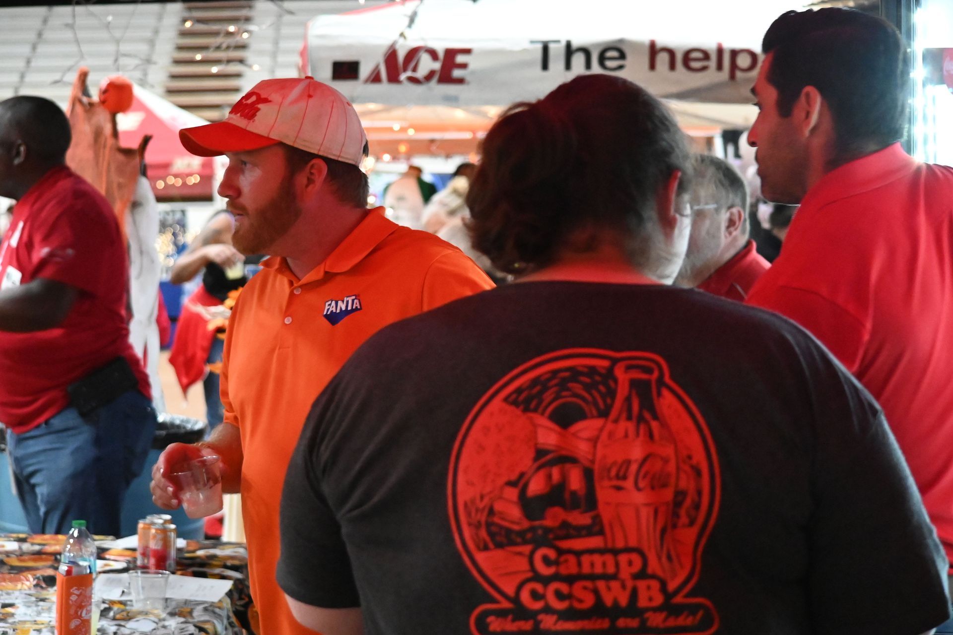 People in red shirts and one in orange chat at a camp booth under a canopy