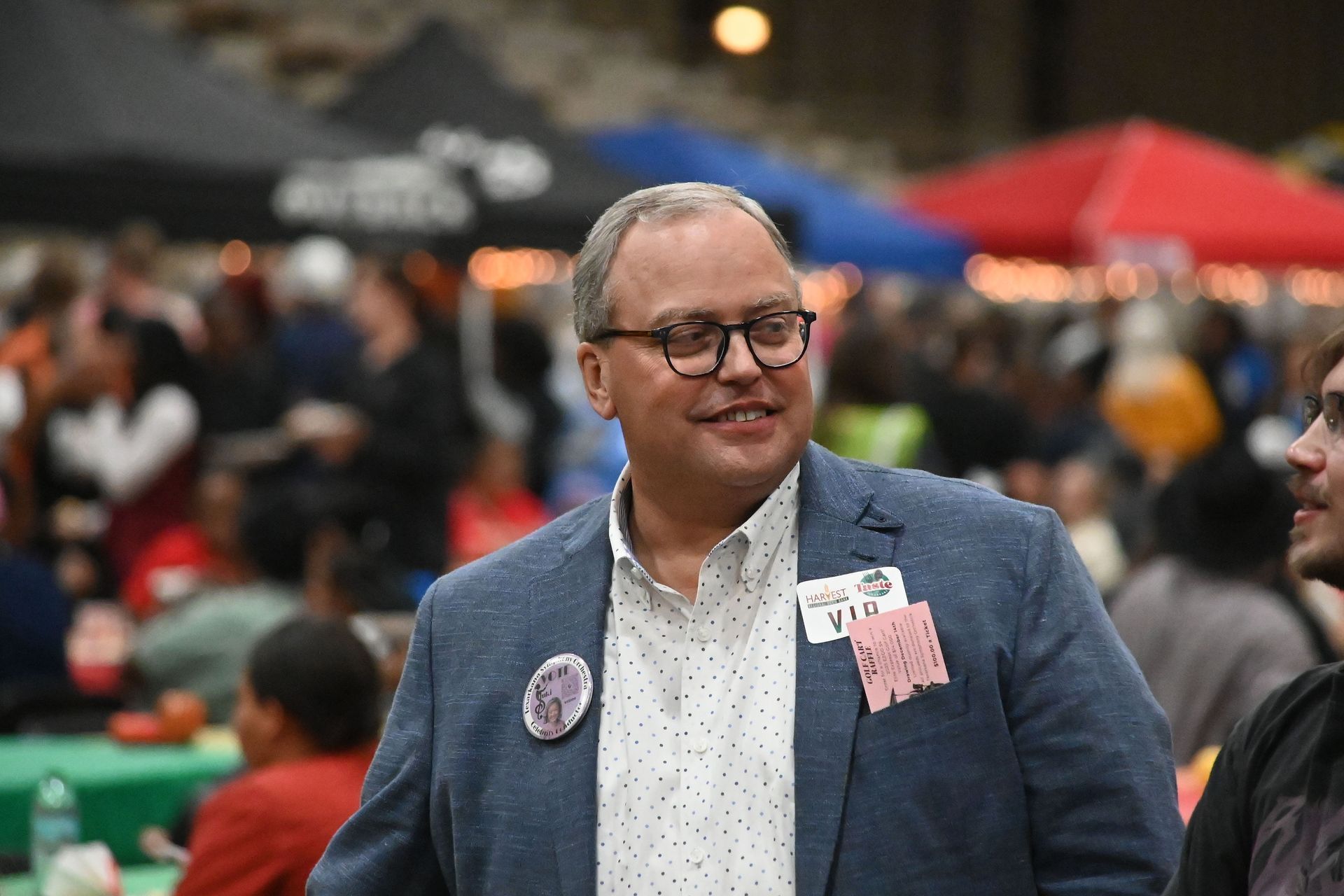 Man in a blue blazer and glasses at a lively outdoor market, with colorful tents blurred behind him