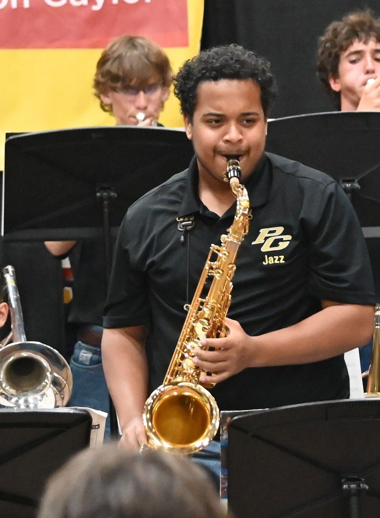Student playing saxophone in a band performance, wearing a black shirt with a brass saxophone and music stands behind him