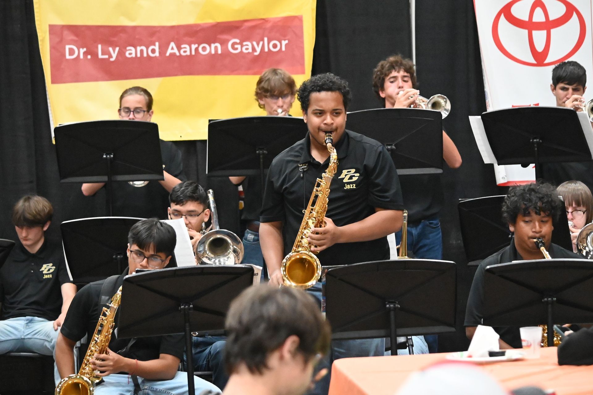 School jazz band performing, with a saxophonist in center and music stands in front of a banner.