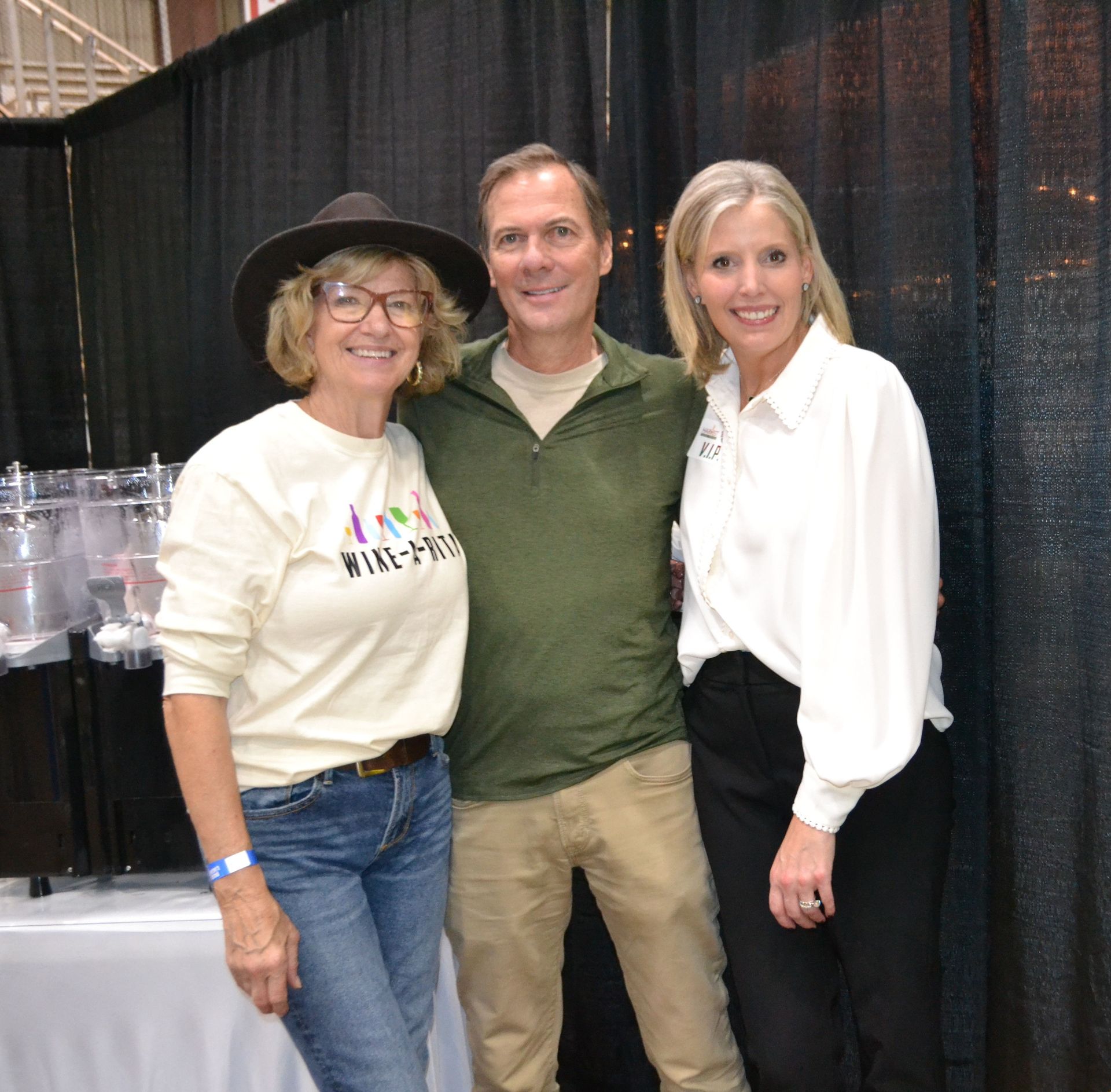 Three people smiling at an indoor event booth with black curtains and tables behind them.