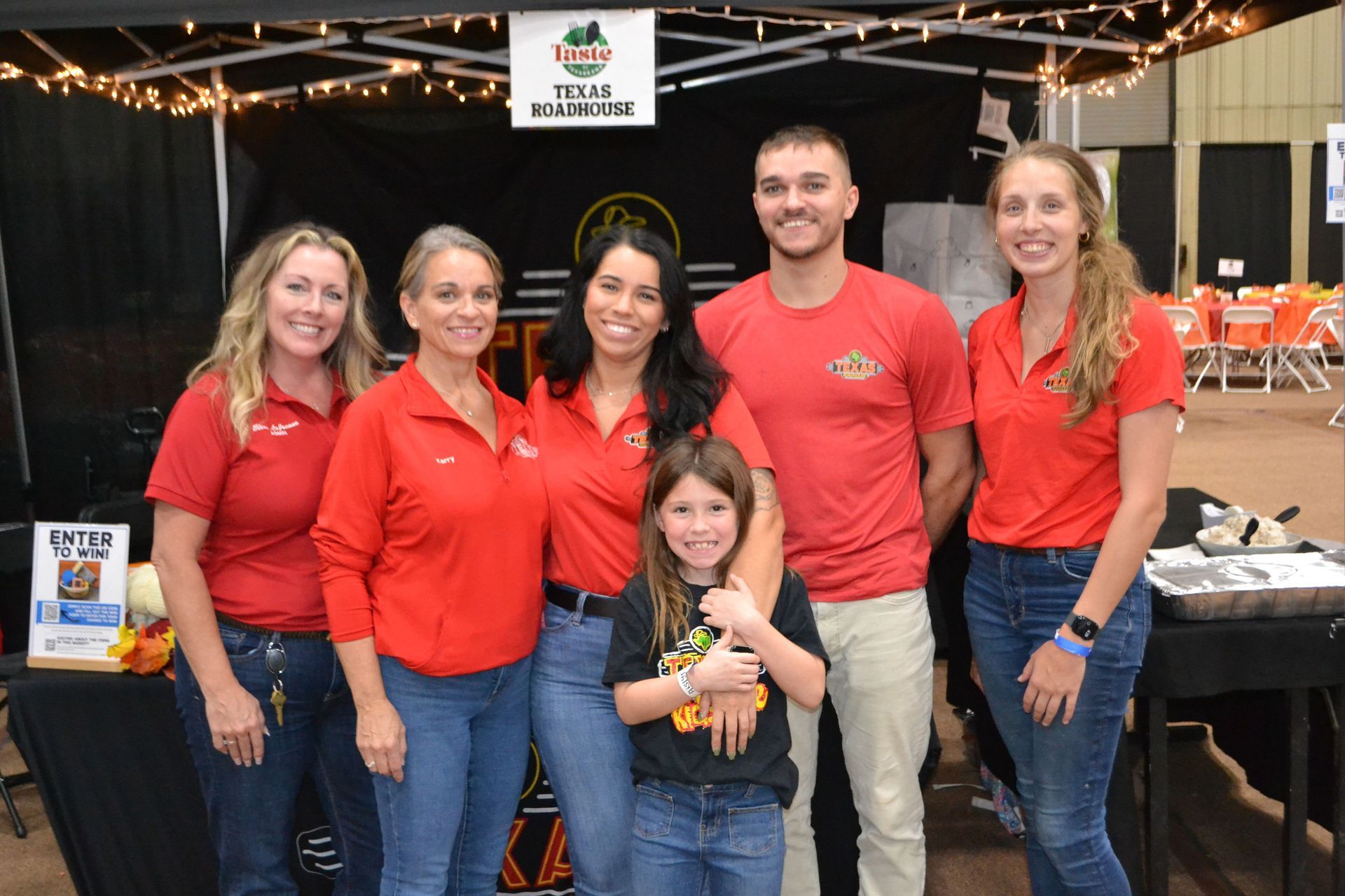 Group of five people in red shirts posing at an indoor event booth with string lights and tables behind them