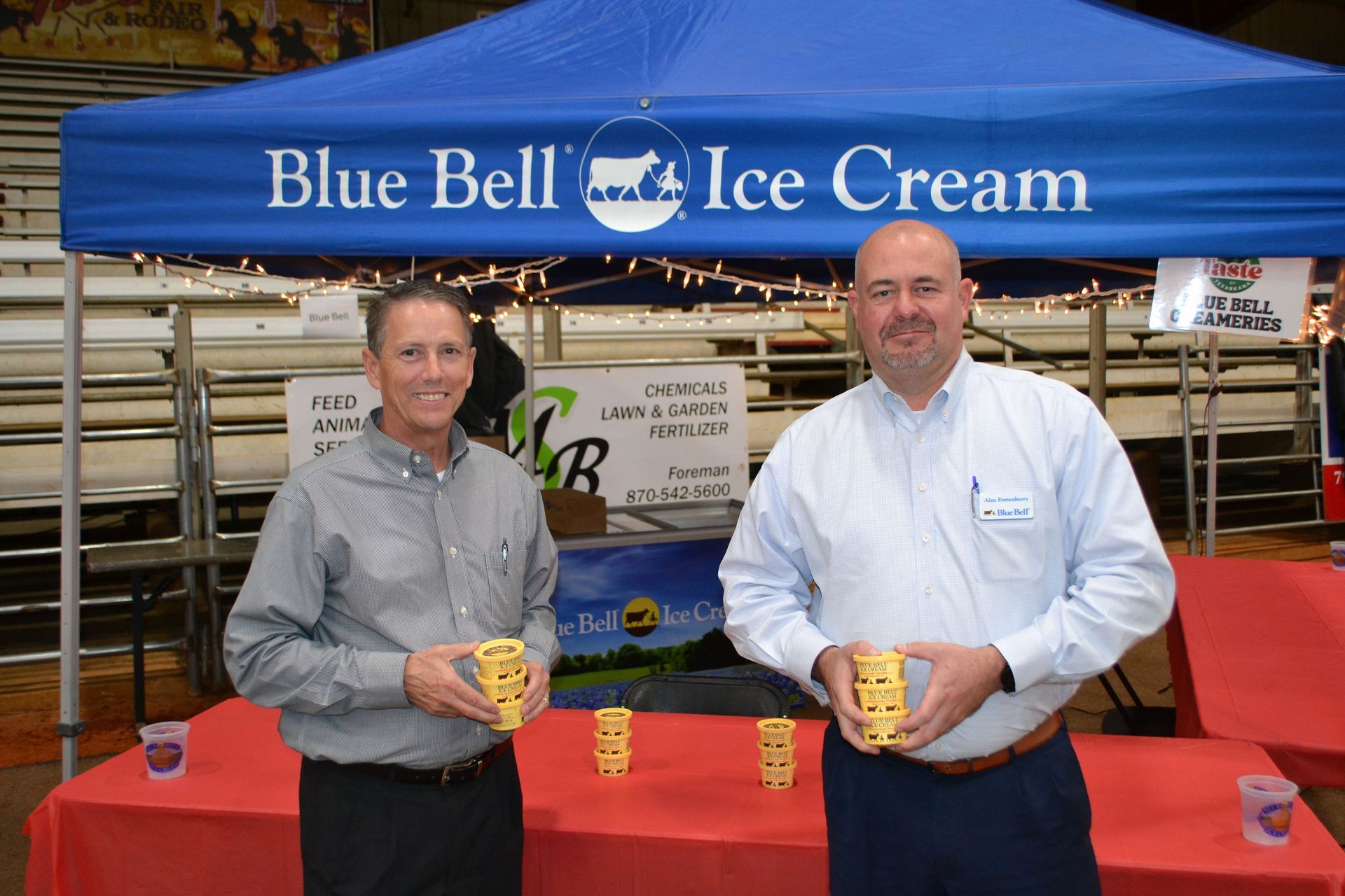 Two men holding Blue Bell ice cream cups at a promotional booth under a blue canopy.