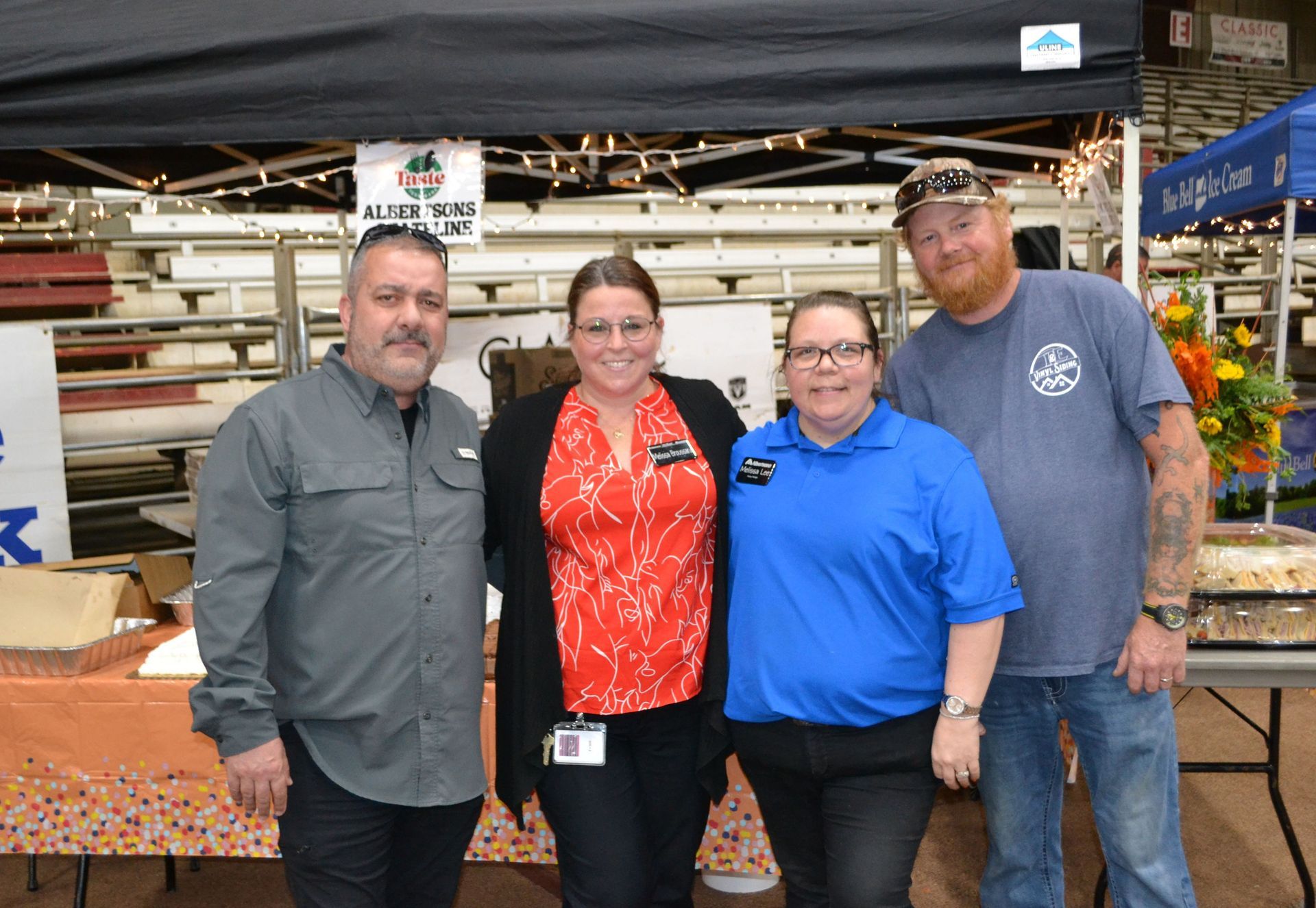 Four people standing together indoors at a market stall, smiling at the camera.