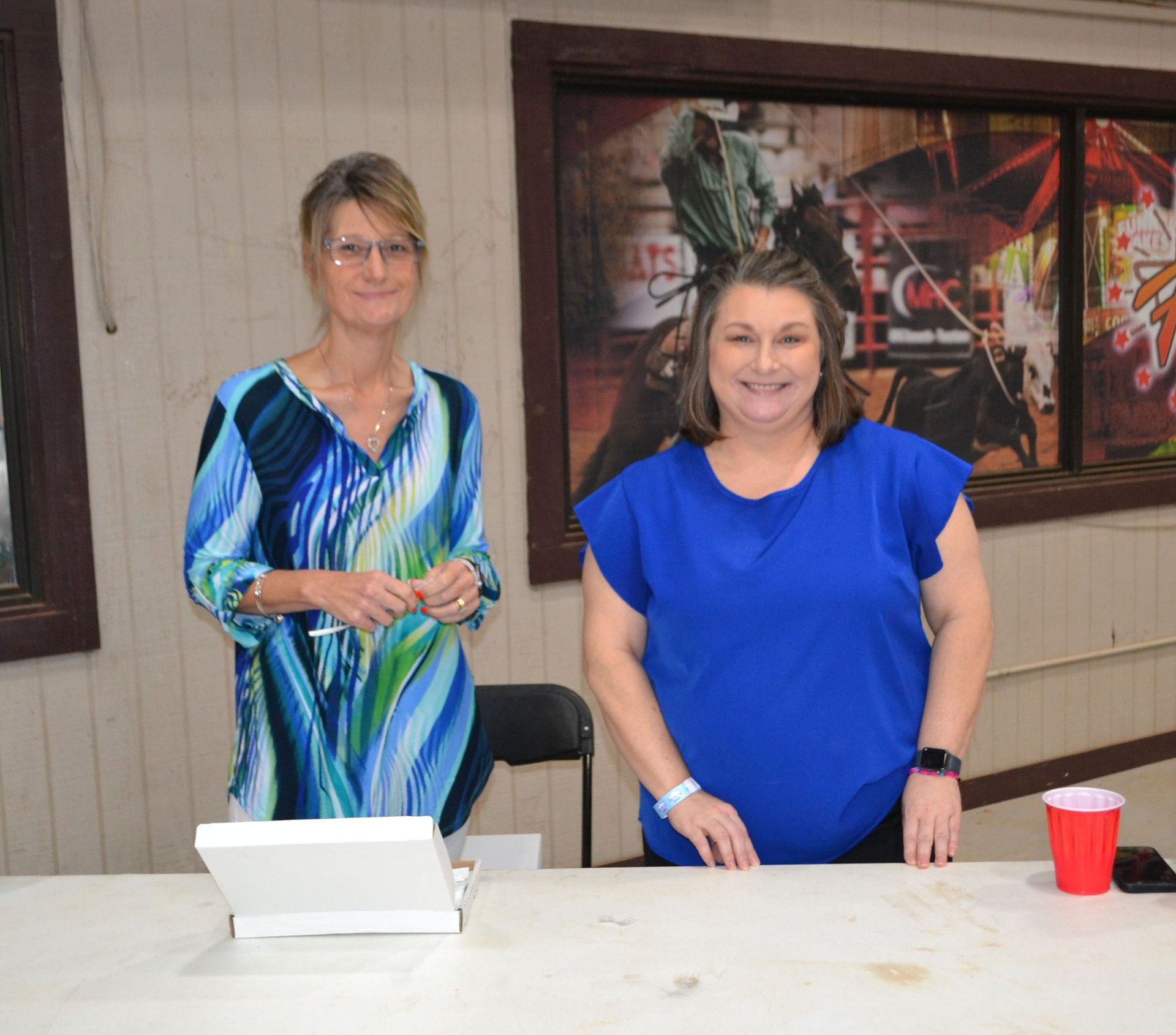 Two women stand behind a table, smiling at an indoor event with a framed wall photo behind them.
