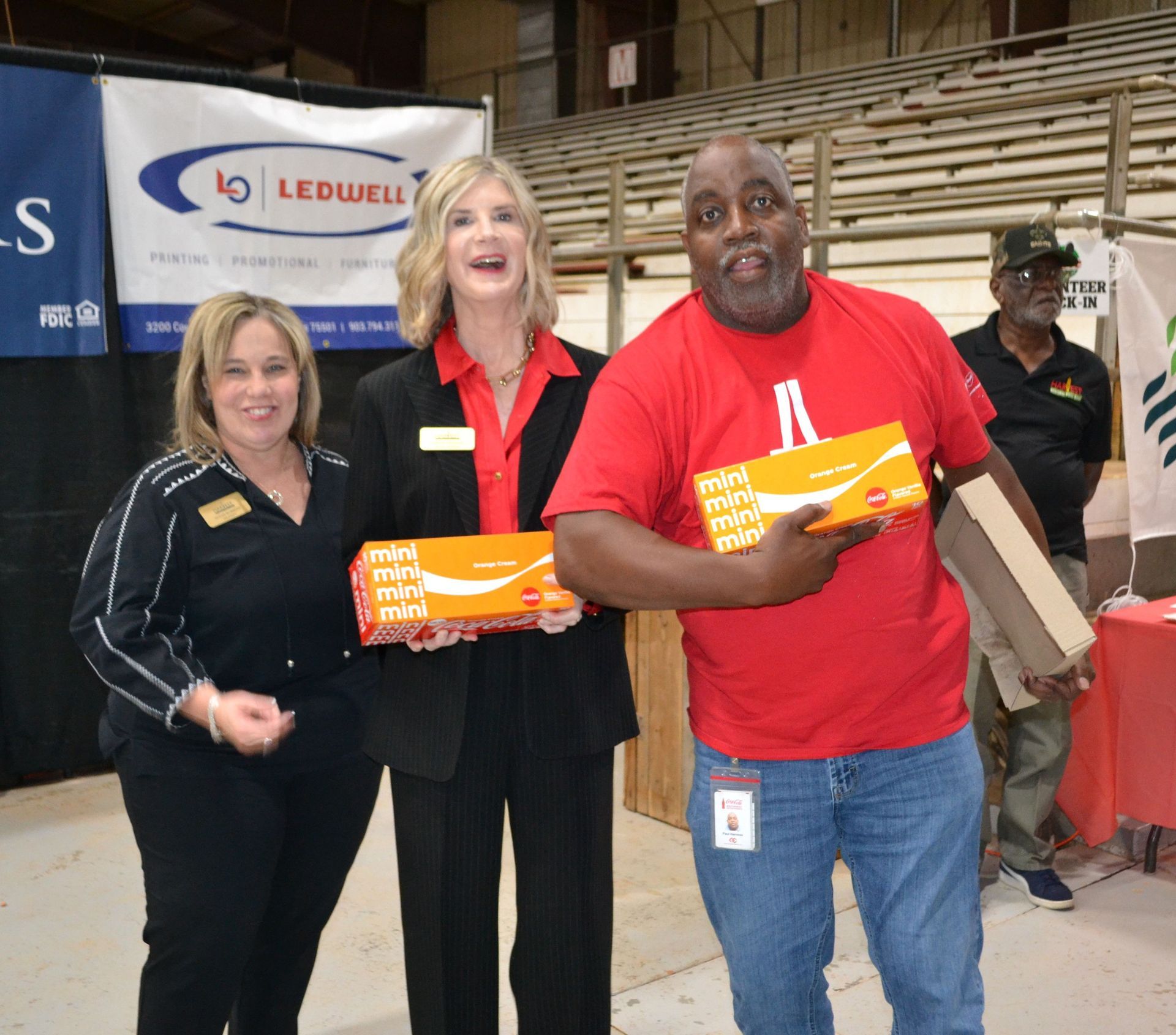 Three volunteers holding orange donation boxes at an indoor event, with bleachers and a Ledwell sign in the background