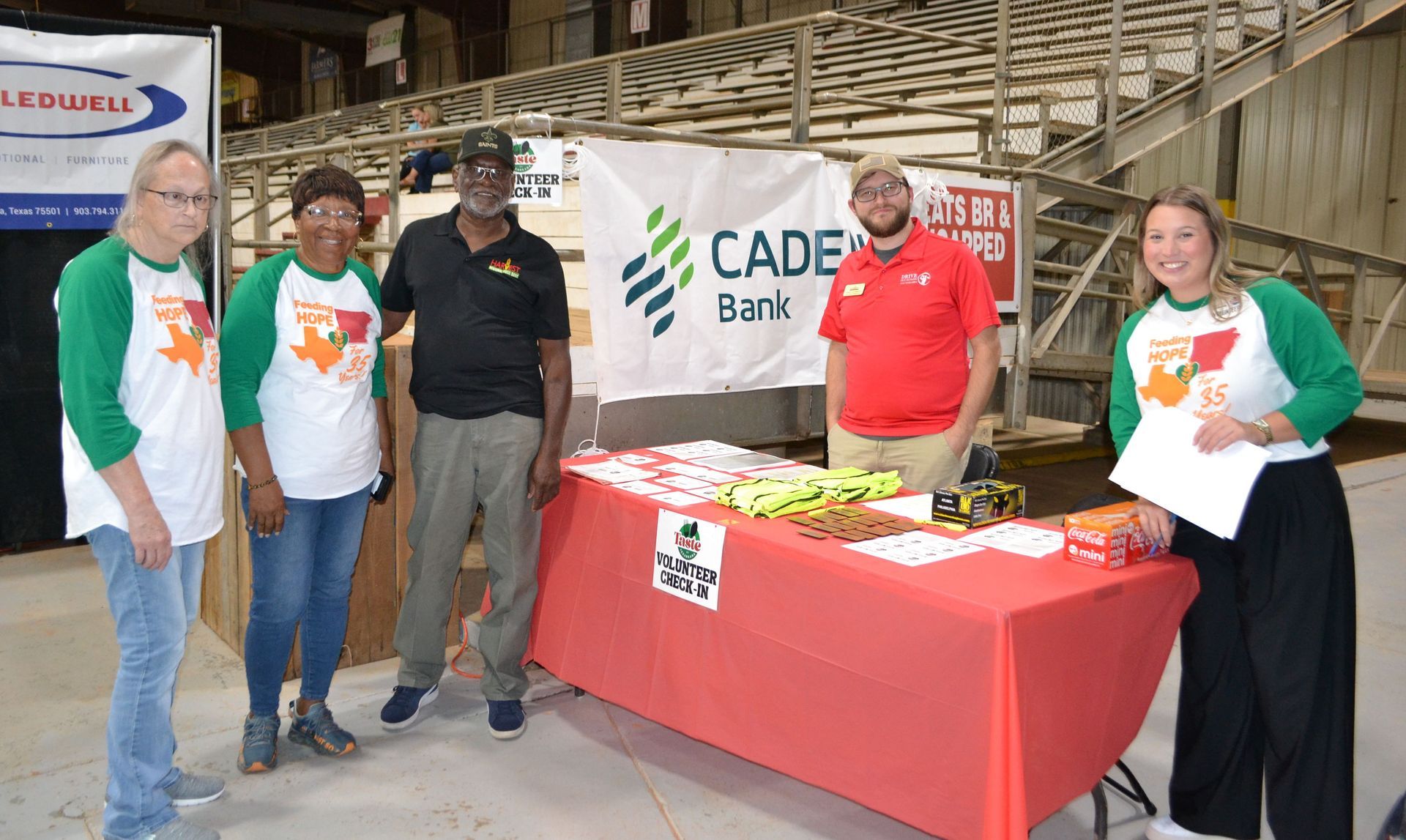 Five people stand behind a red table at a CADE Bank event booth with brochures and snacks.