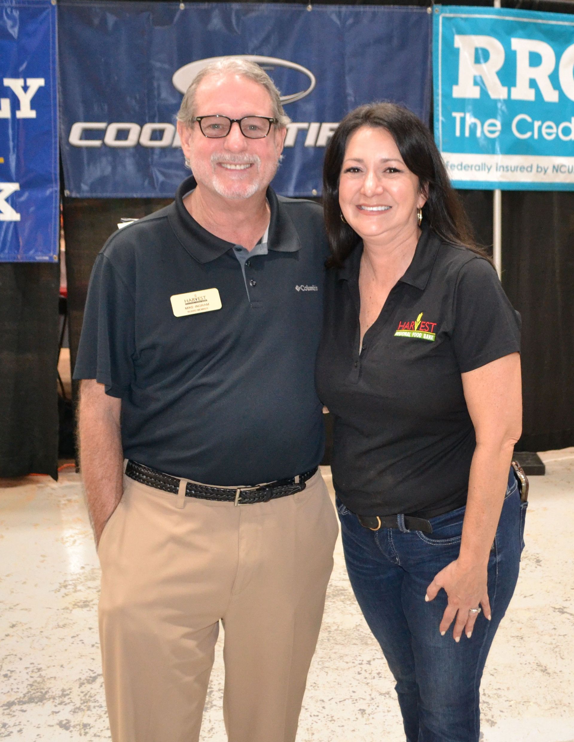 Two smiling people standing together at an indoor event booth, wearing black shirts and khaki pants/jeans.