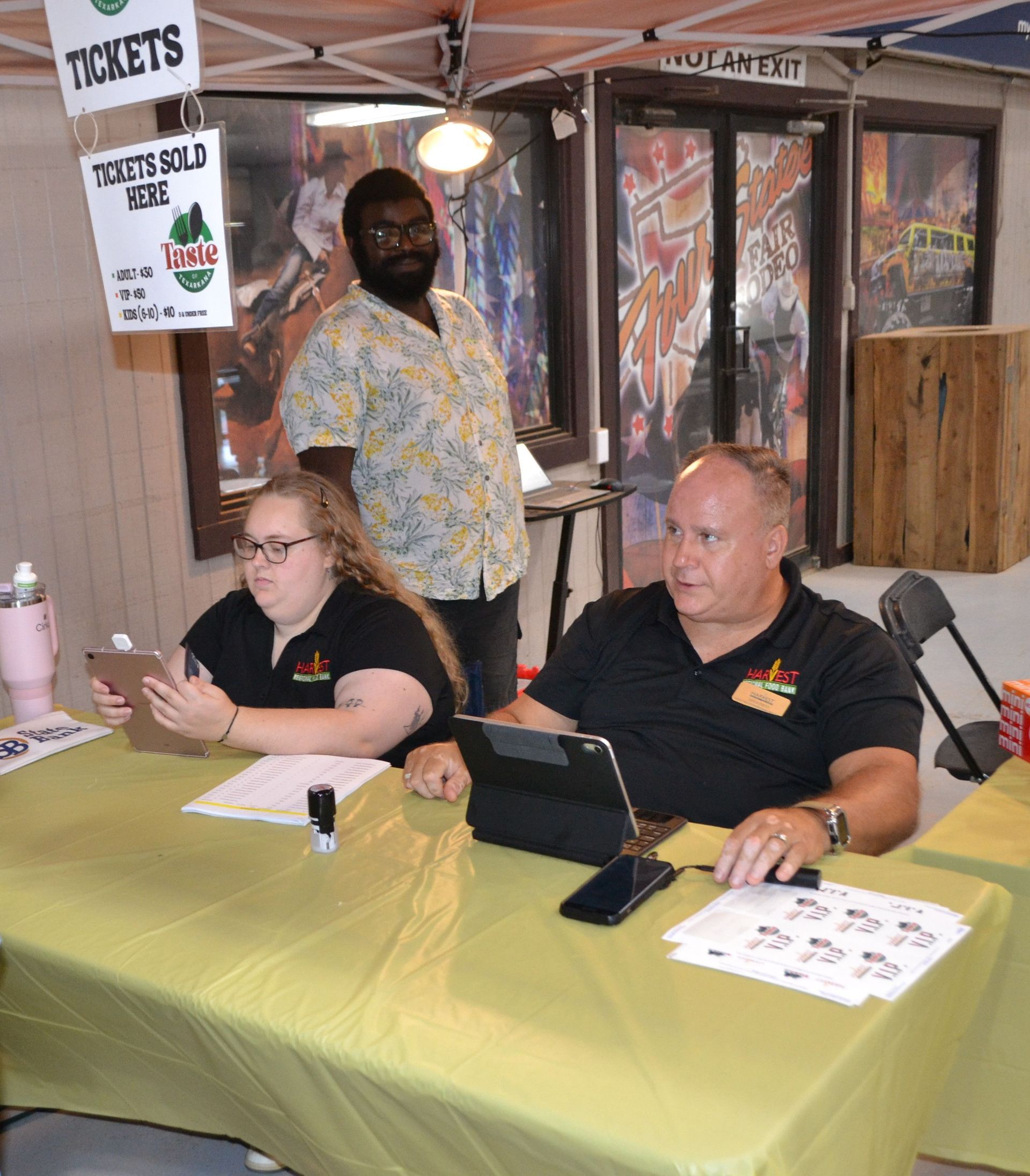 Two people at a registration table under a “Tickets” sign, with papers and laptops on the table.