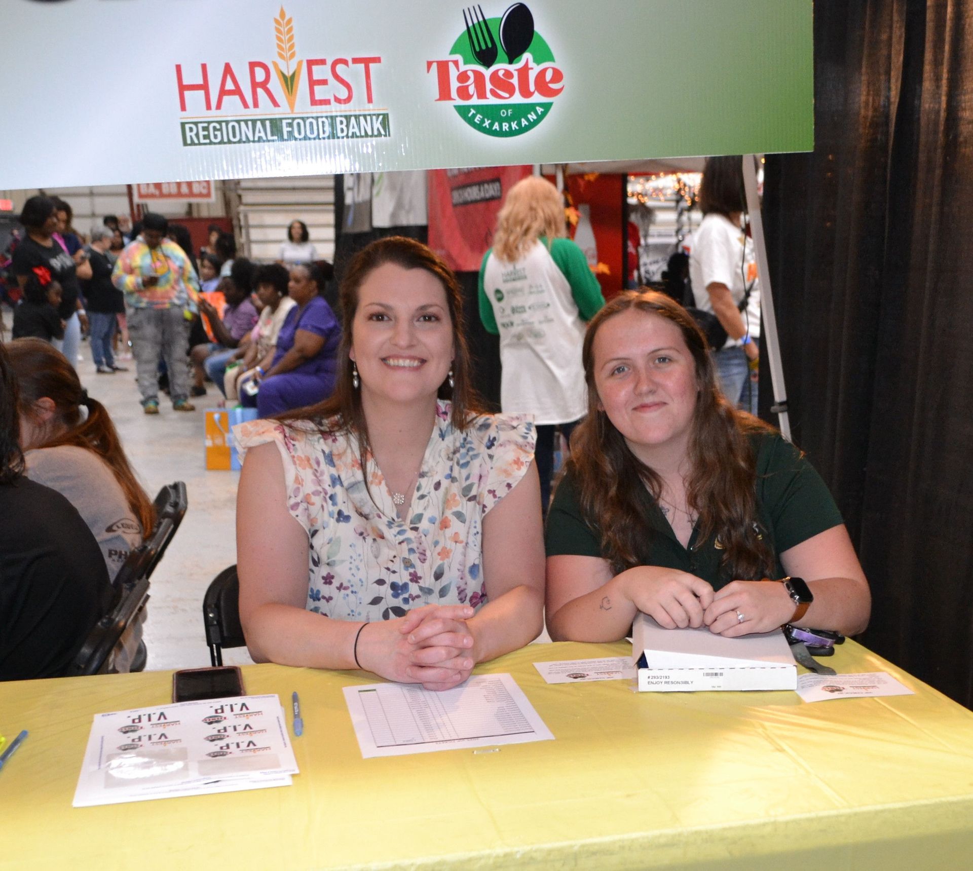 Two women sit behind a yellow table at a Harvest Taste event booth with signs and flyers.