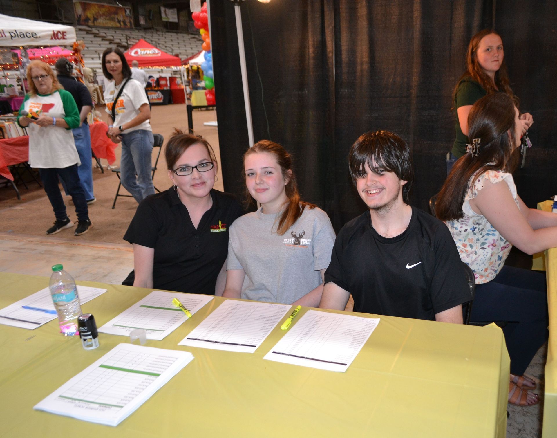 Three people seated at a yellow table with forms, at an indoor event booth.