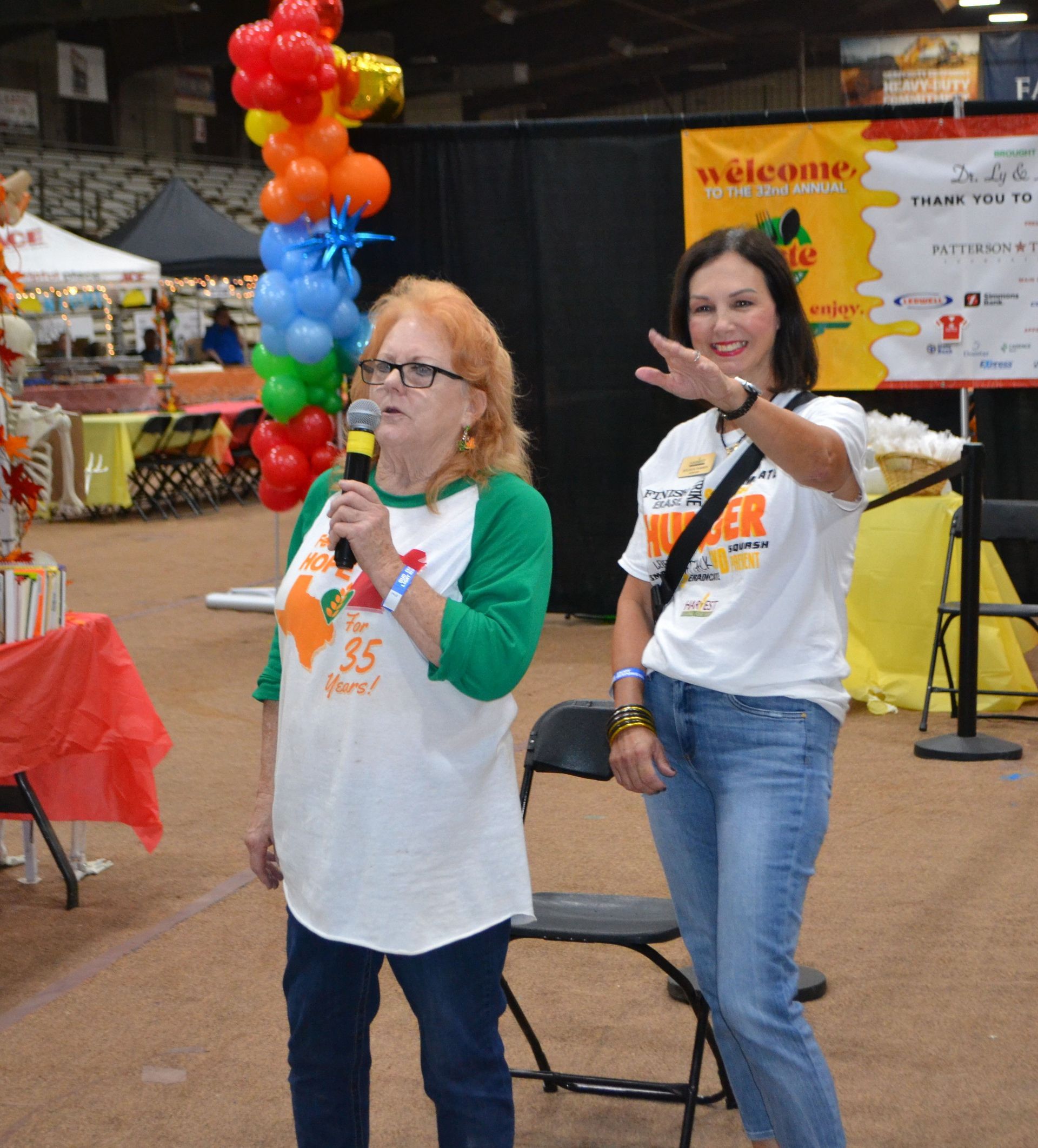 Two women in white shirts at a fair, one speaking into a microphone and one gesturing beside balloon decorations.