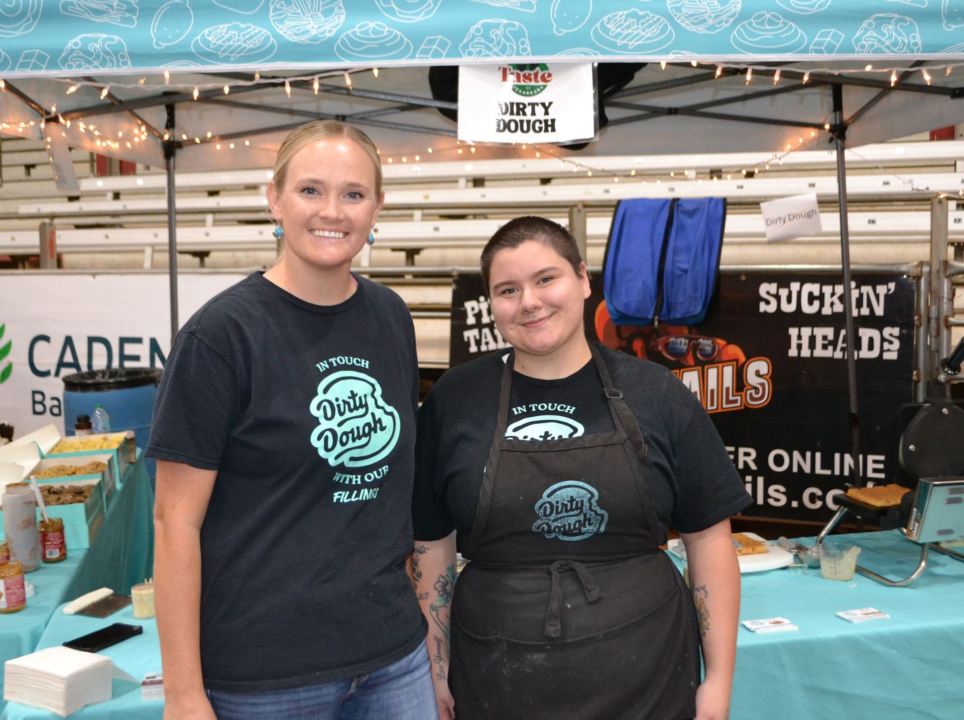 Two people smiling at a vendor booth with T-shirts and a sign reading “Dirty Book”