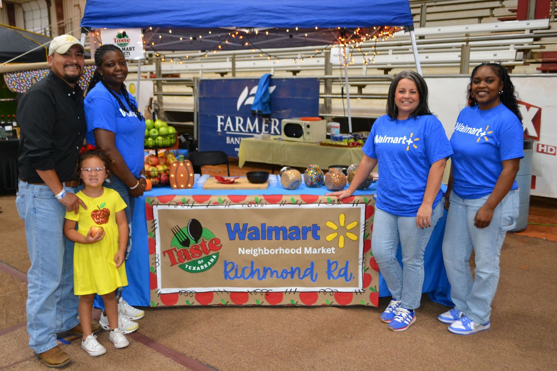 People posing at a Walmart Community garden booth with fresh produce and a banner at an outdoor market.