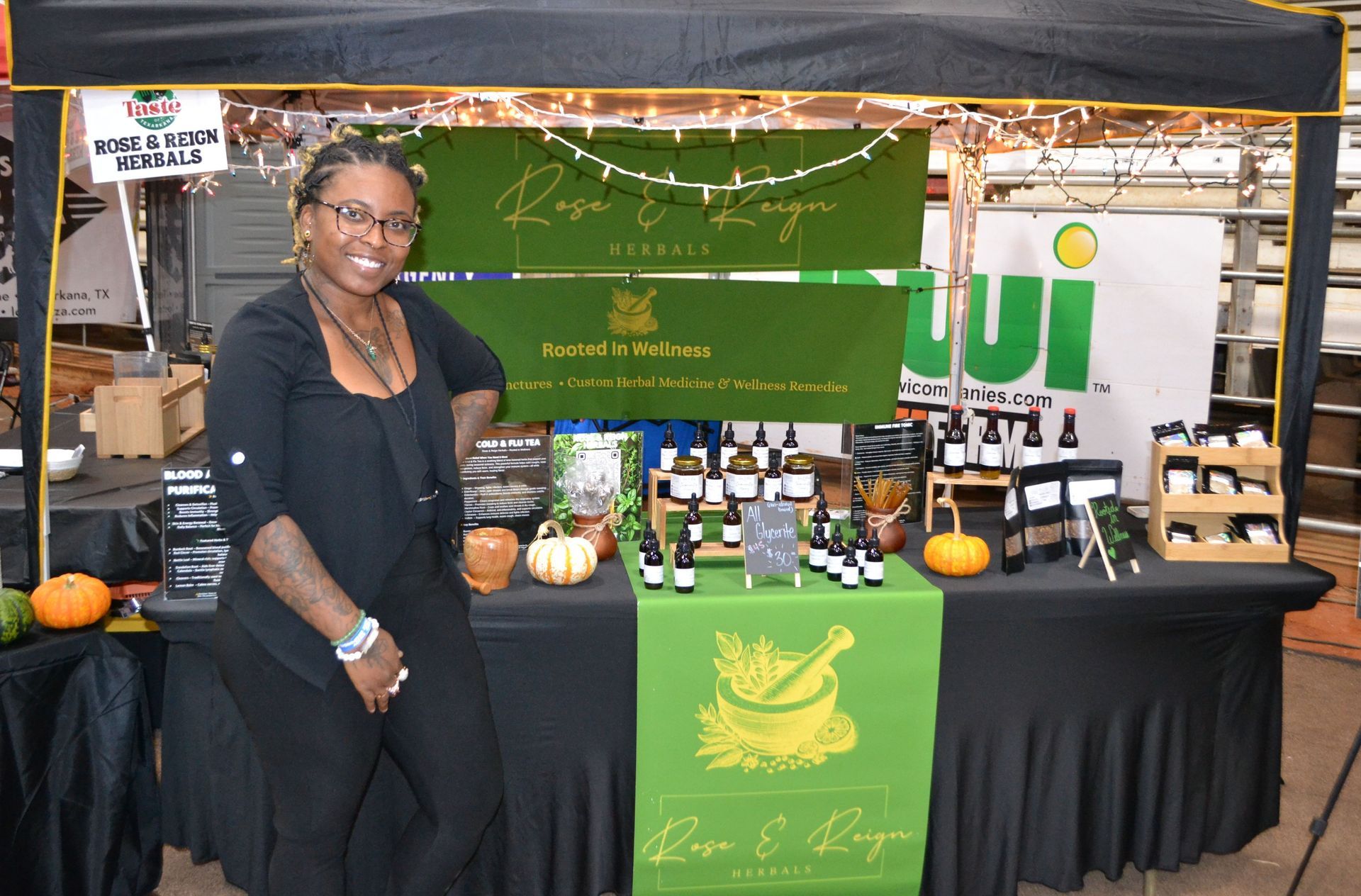 Woman standing at a market booth with green branding, bottles, and pumpkins on display