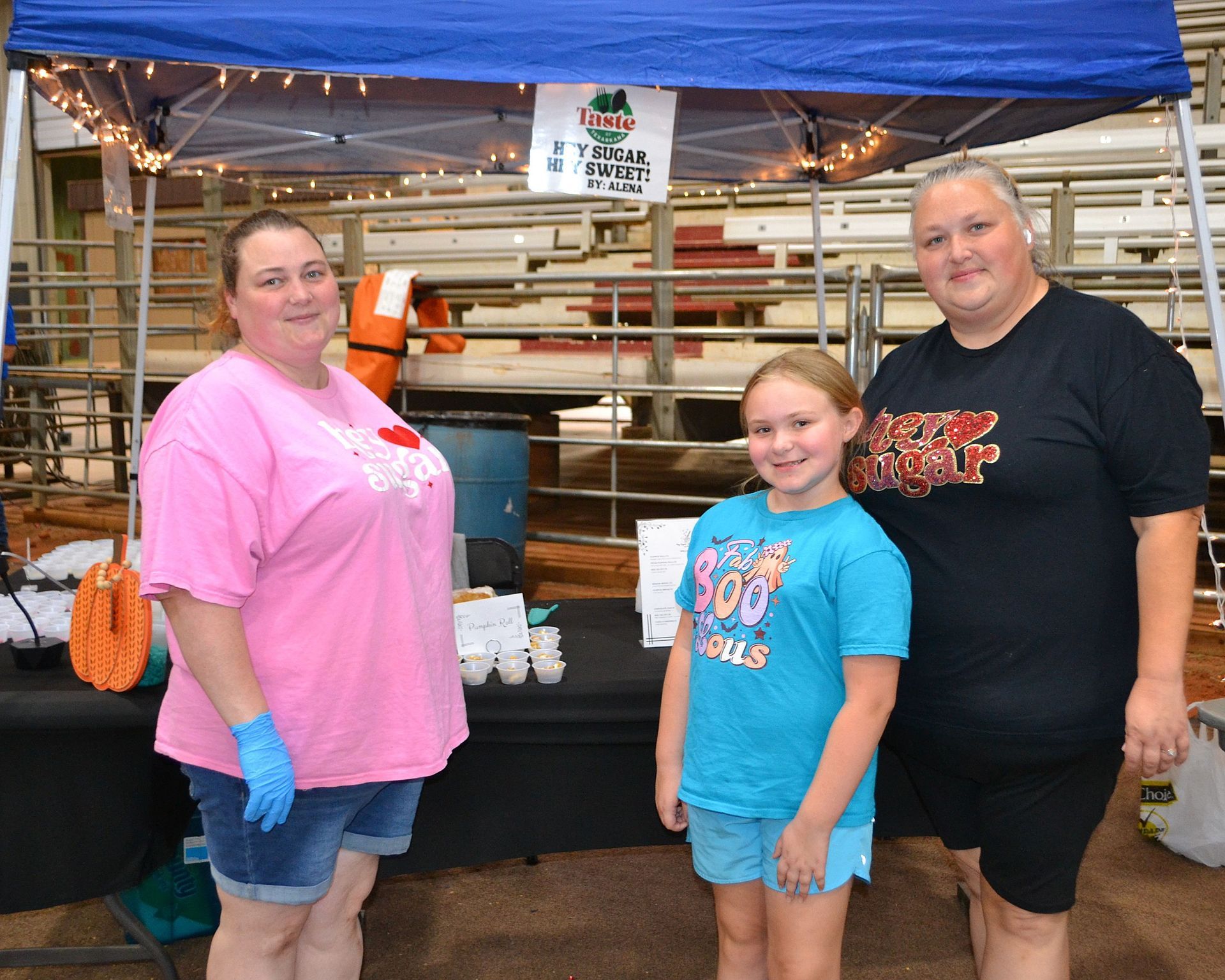 Three people stand at a market booth under a blue canopy with shelves and folded items behind them.