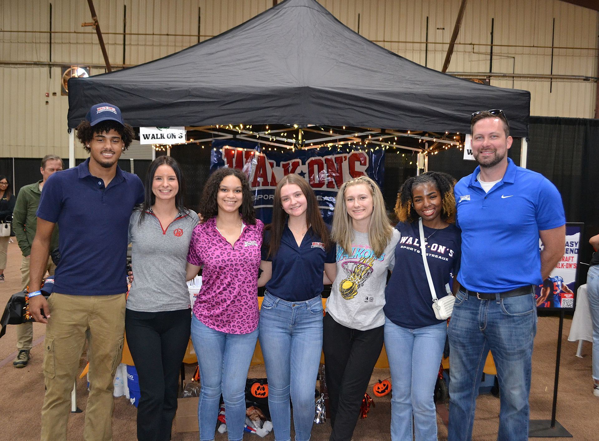 Group of seven smiling people standing in front of a tented booth at an indoor event