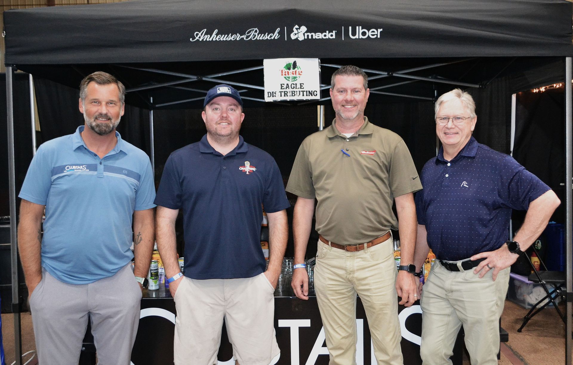 Four men posing in front of a branded tent at an event booth.