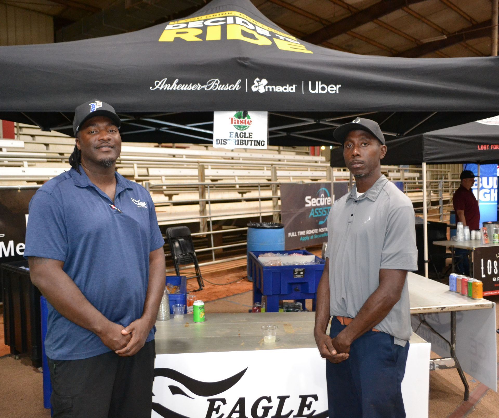 Two men standing behind a branded booth under a black canopy at an indoor event.