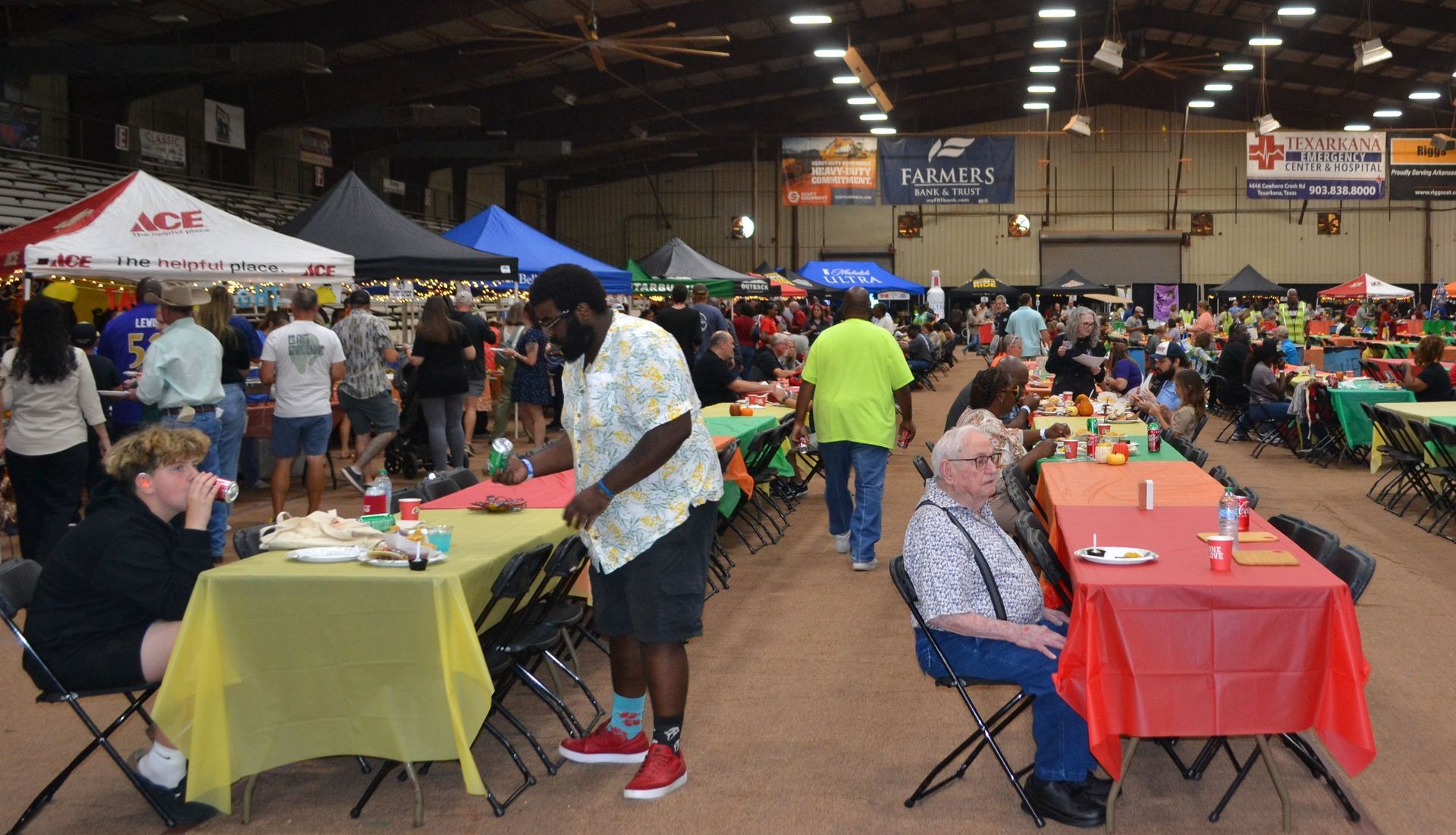 Crowded indoor market with vendors and shoppers at tables covered in red, yellow, and green tablecloths.