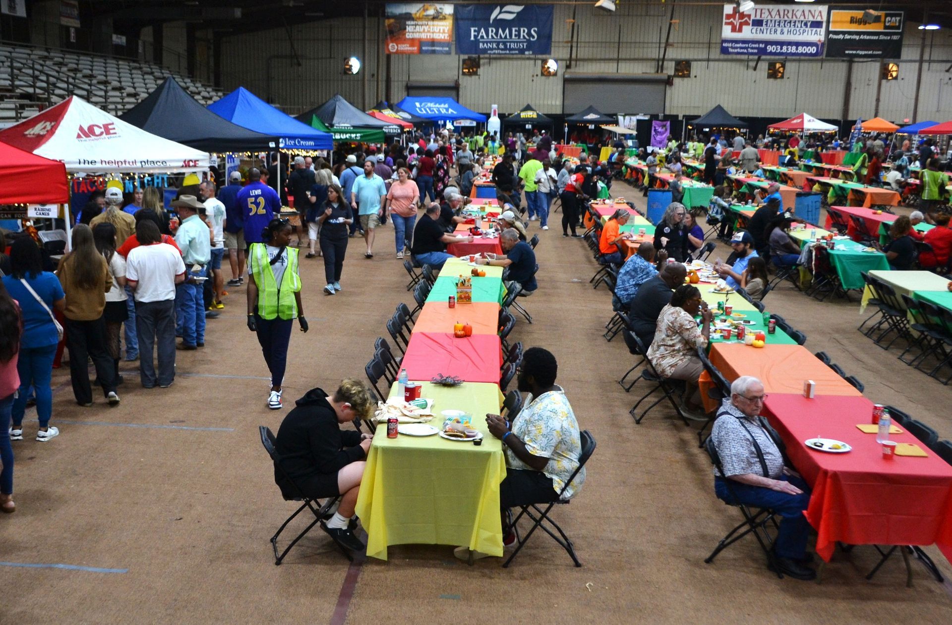 Crowded indoor market or fair with rows of colorful tables and people seated and walking around