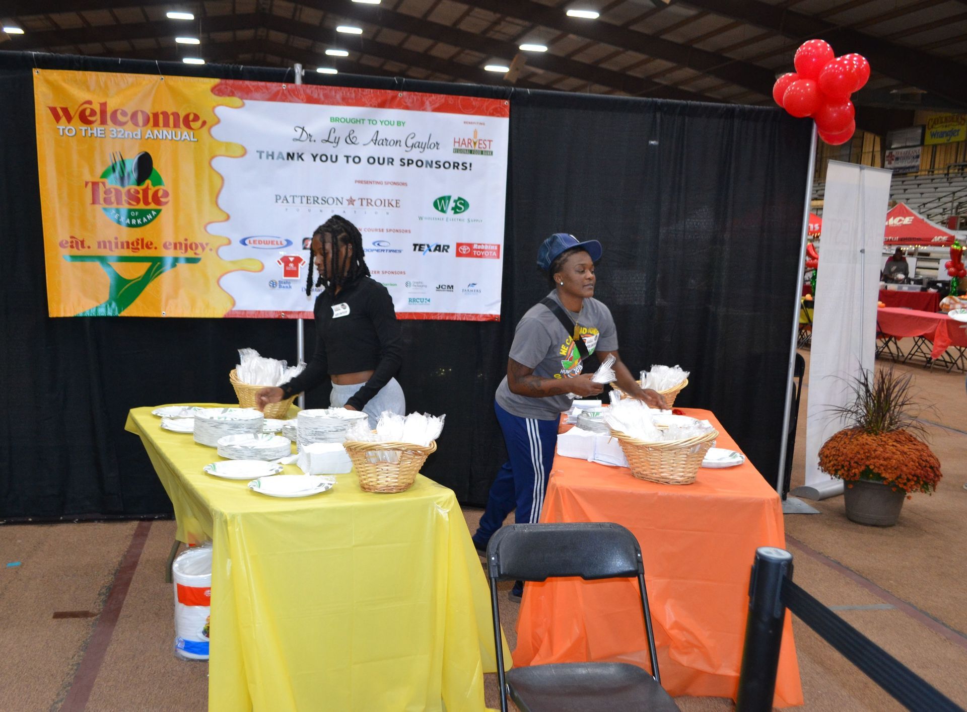 Indoor event booth with colorful tablecloths, banner backdrop, and two people arranging food samples