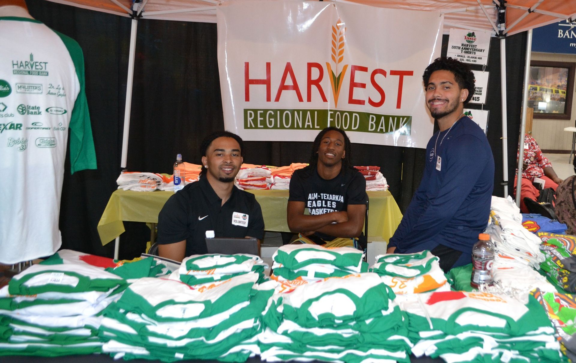 Harvest Regional Food Bank booth with three volunteers behind a table of green-and-white T-shirts and folded clothes