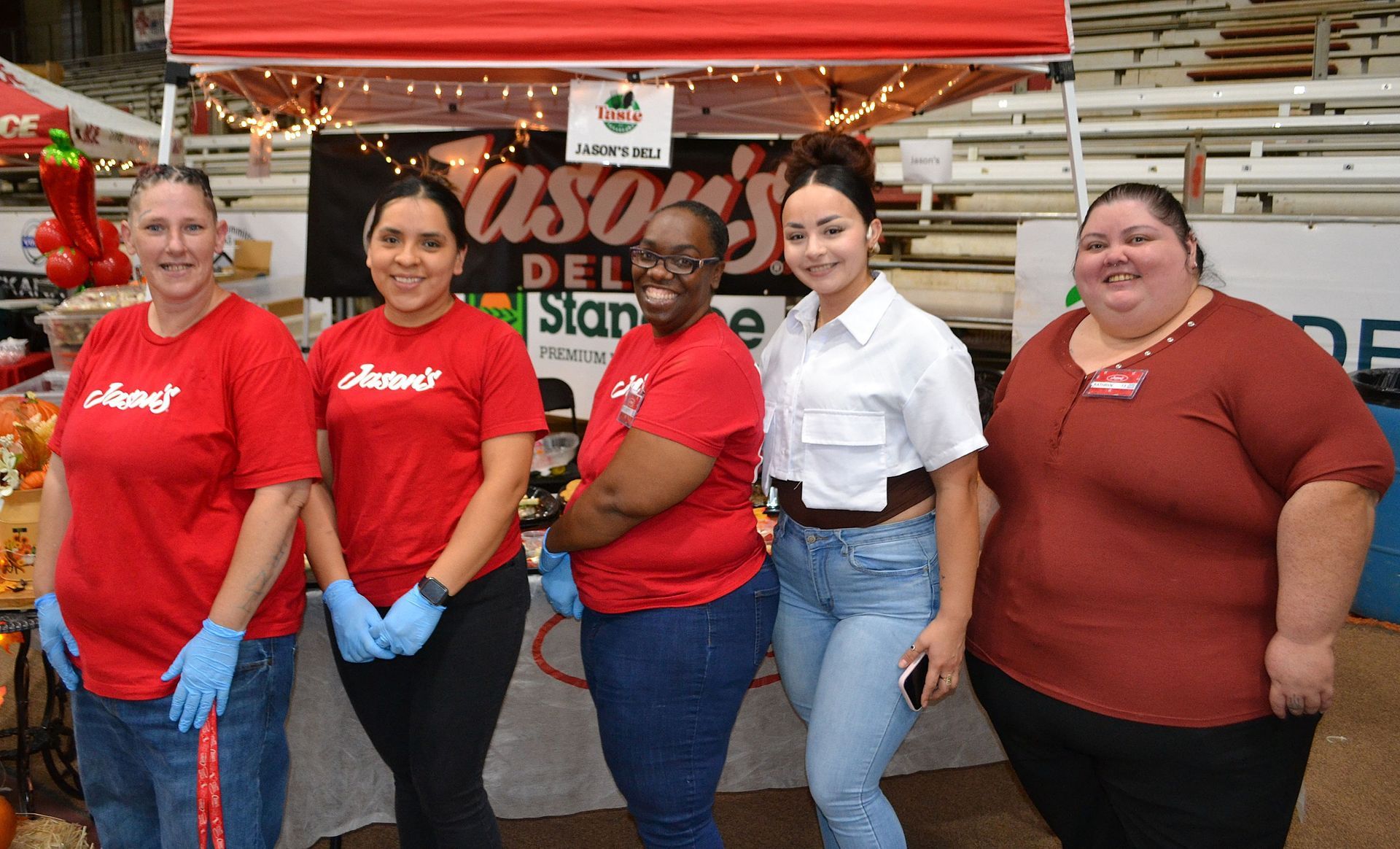 Five people stand in front of a red market stall, two wearing red shirts and one in white.