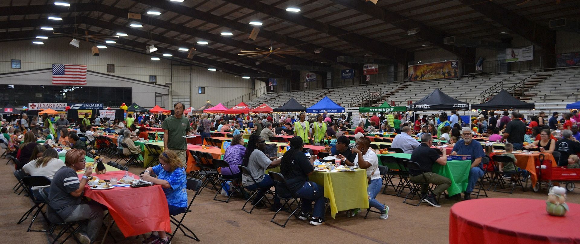 Large indoor banquet hall with many people seated at colorful tables under a high roof