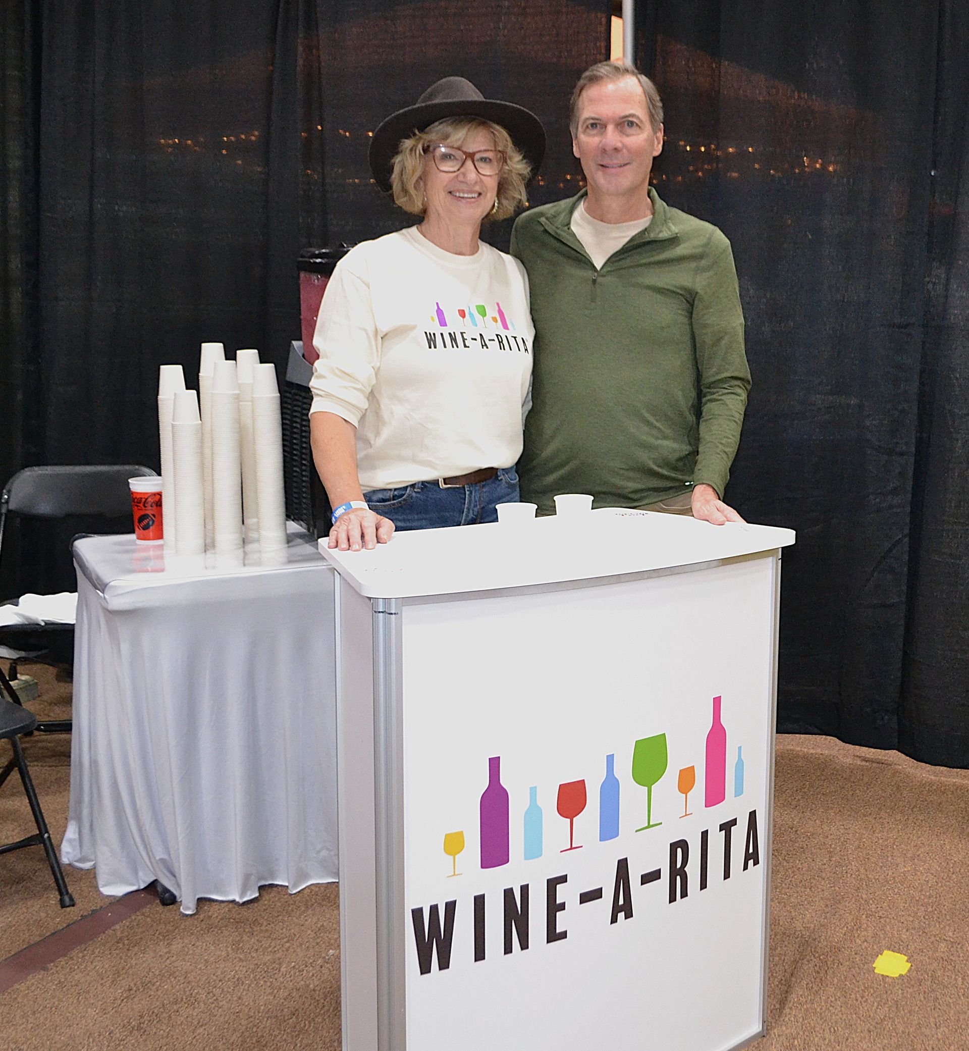 Two people behind a Wine-a-Rita booth with cups and a white tablecloth at an indoor event