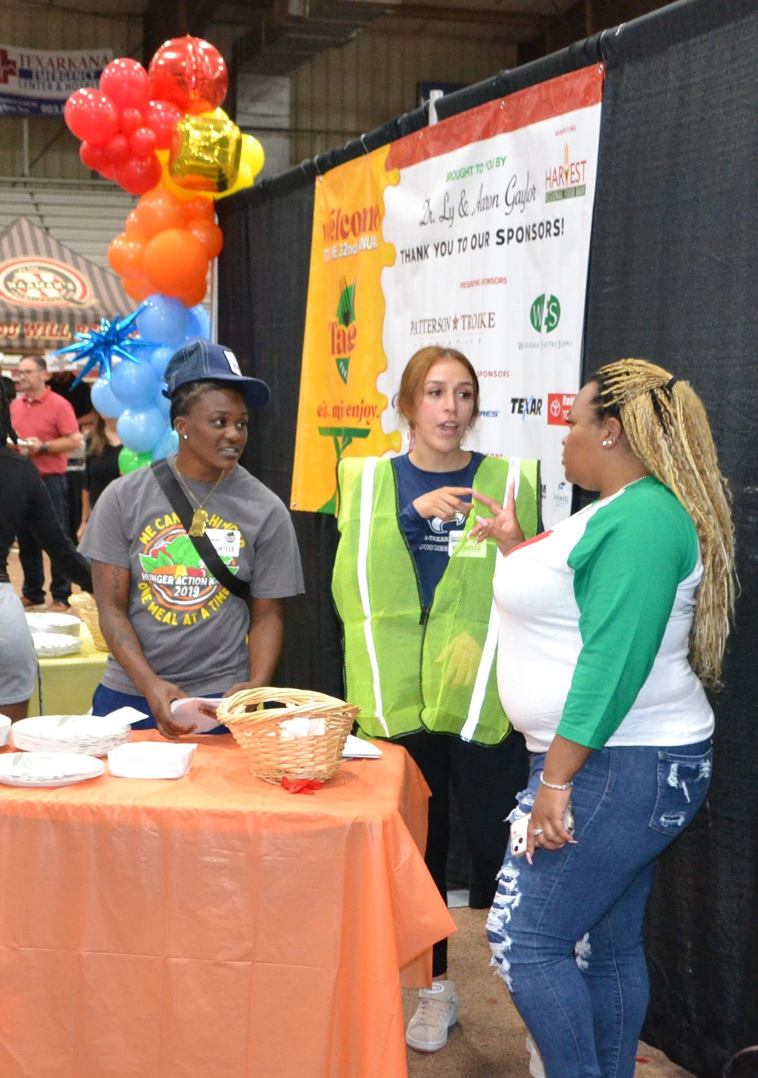 Three people chatting at an orange-covered booth with balloons and a banner at an event.