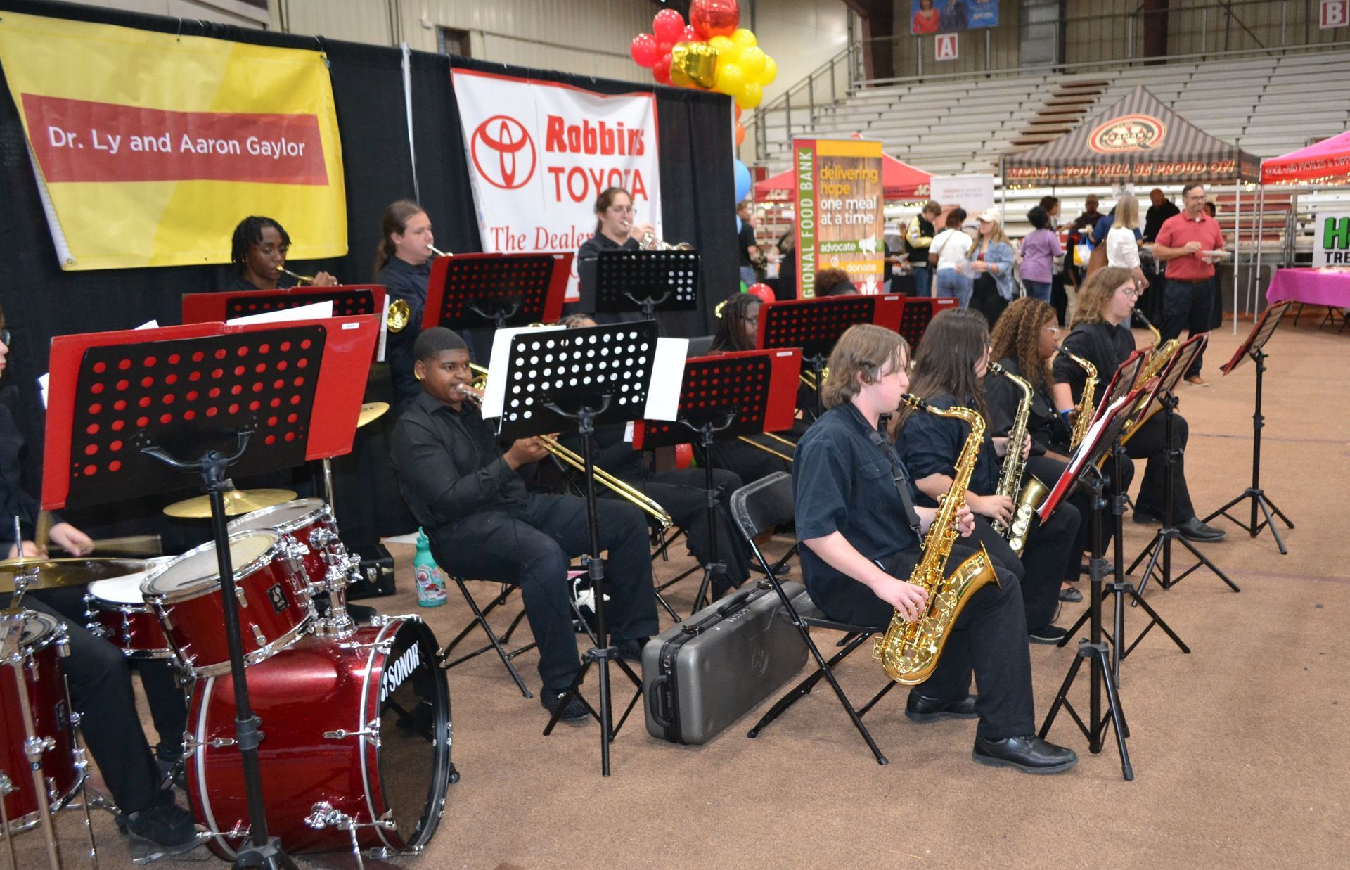 Jazz band performing indoors with drums, saxophones, and music stands at a market or event venue