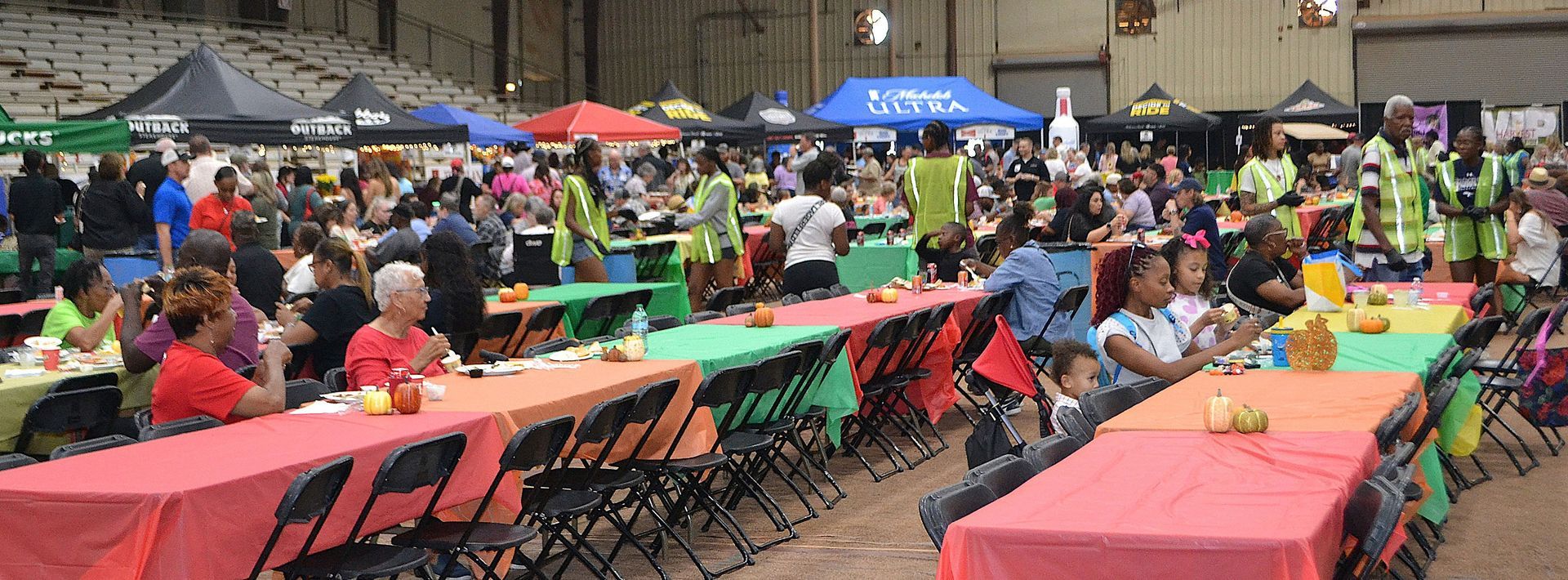 Outdoor community gathering with colorful tables and tents, people seated and socializing in a plaza