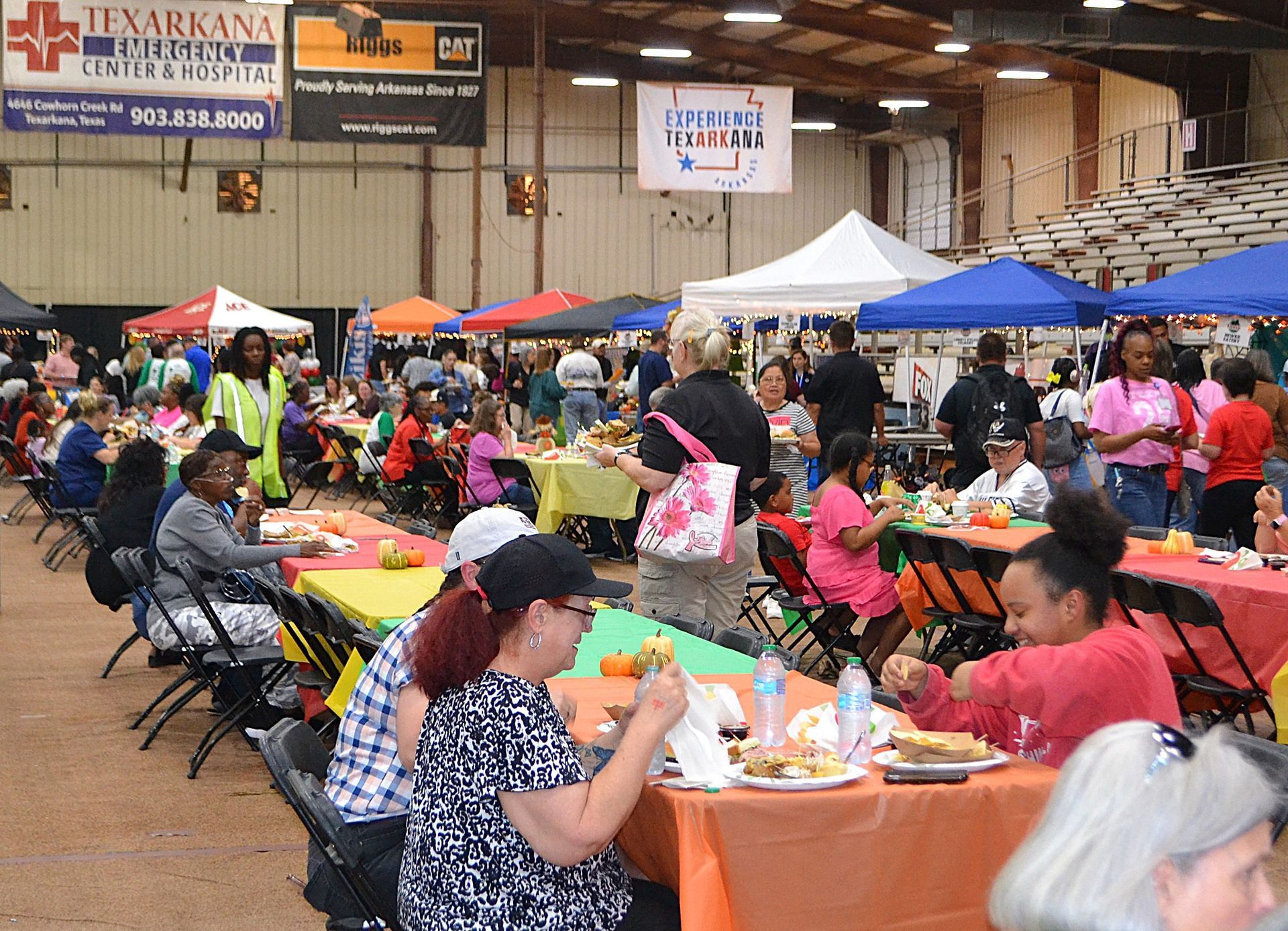 Crowded indoor community fair with people seated at long tables and colorful vendor tents in the background