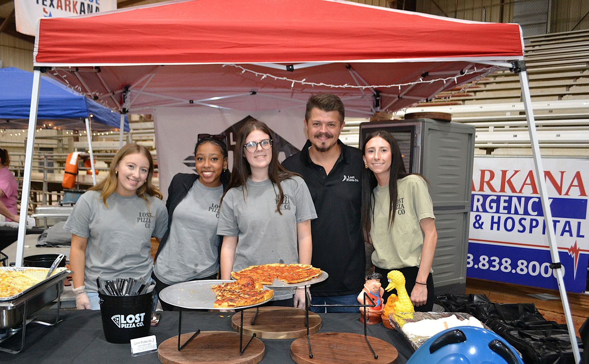 Five people pose under a red canopy beside trays of pizza at an outdoor food stand.