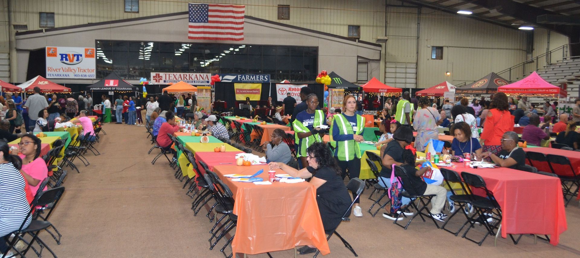 Indoor community fair with rows of tables, colorful tents, and people seated under an American flag banner
