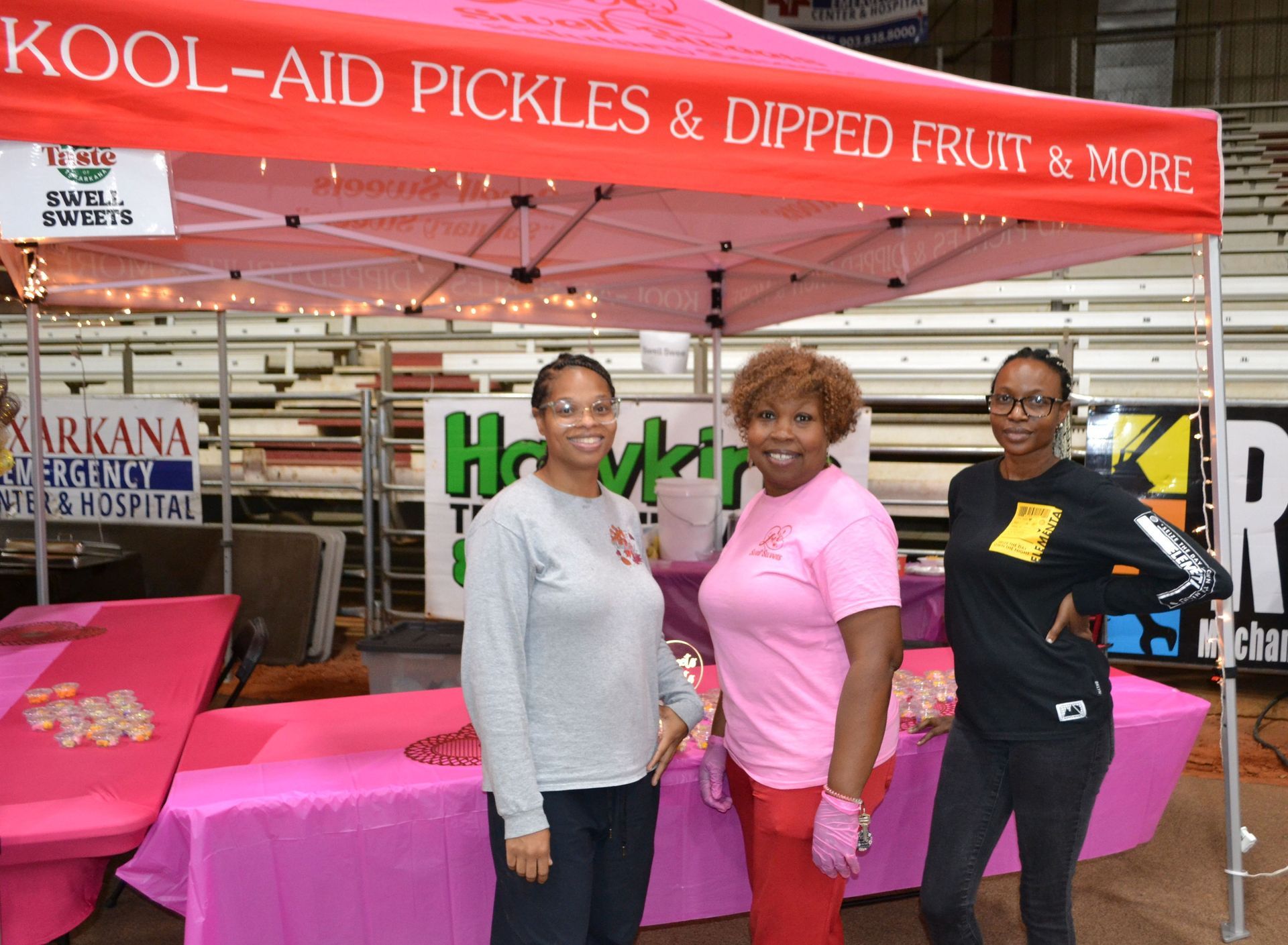Three people posing beside a red Kool-Aid Pickles booth at an indoor market
