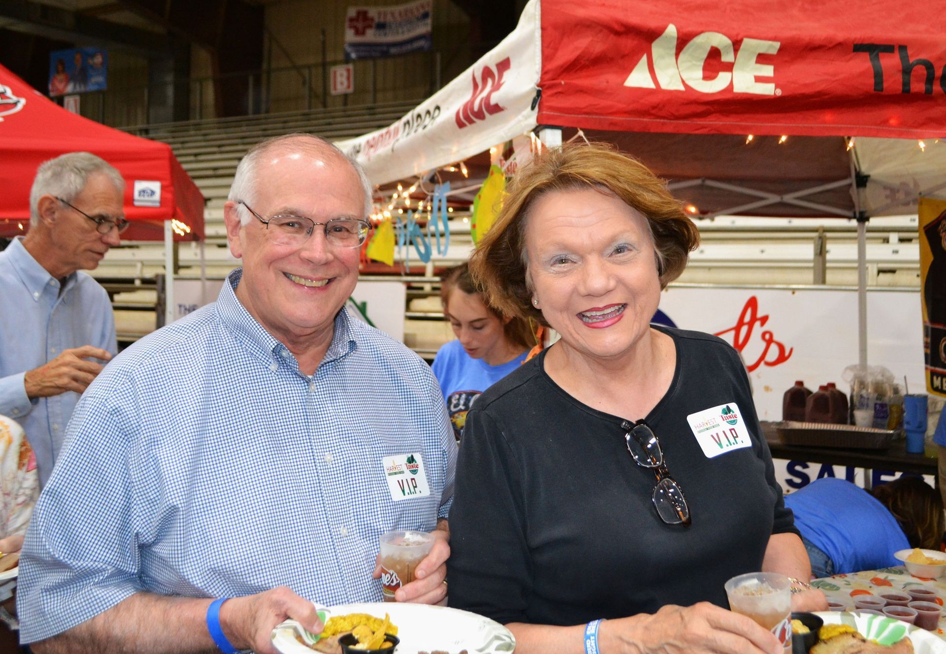 Two smiling people holding plates at a busy indoor food event under an ACE canopy