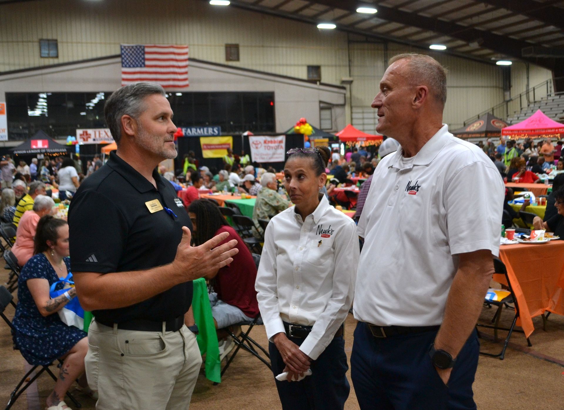 Three people talking at an indoor community event with tables, tents, and an American flag in the background