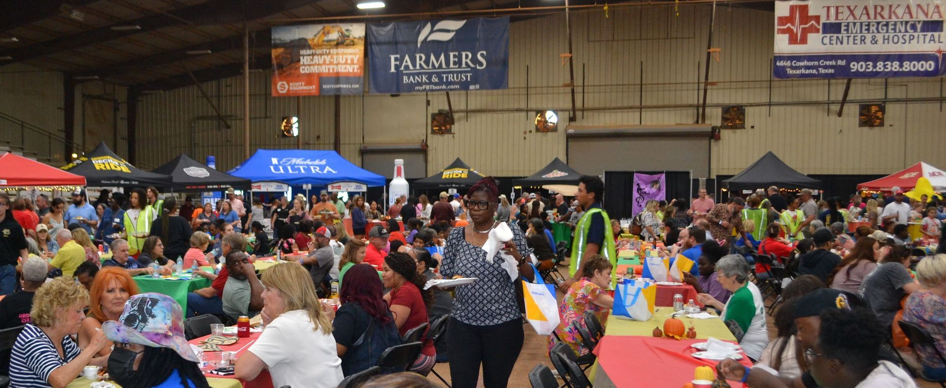 Crowded indoor food fair with colorful tables, people seated and standing, and vendor signs in the background