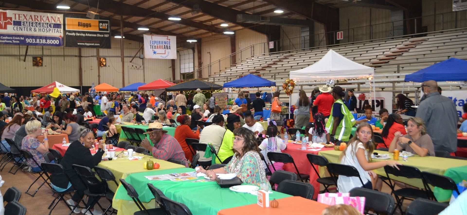 Crowded indoor event with people seated at tables under colorful umbrellas and banners in a large hall